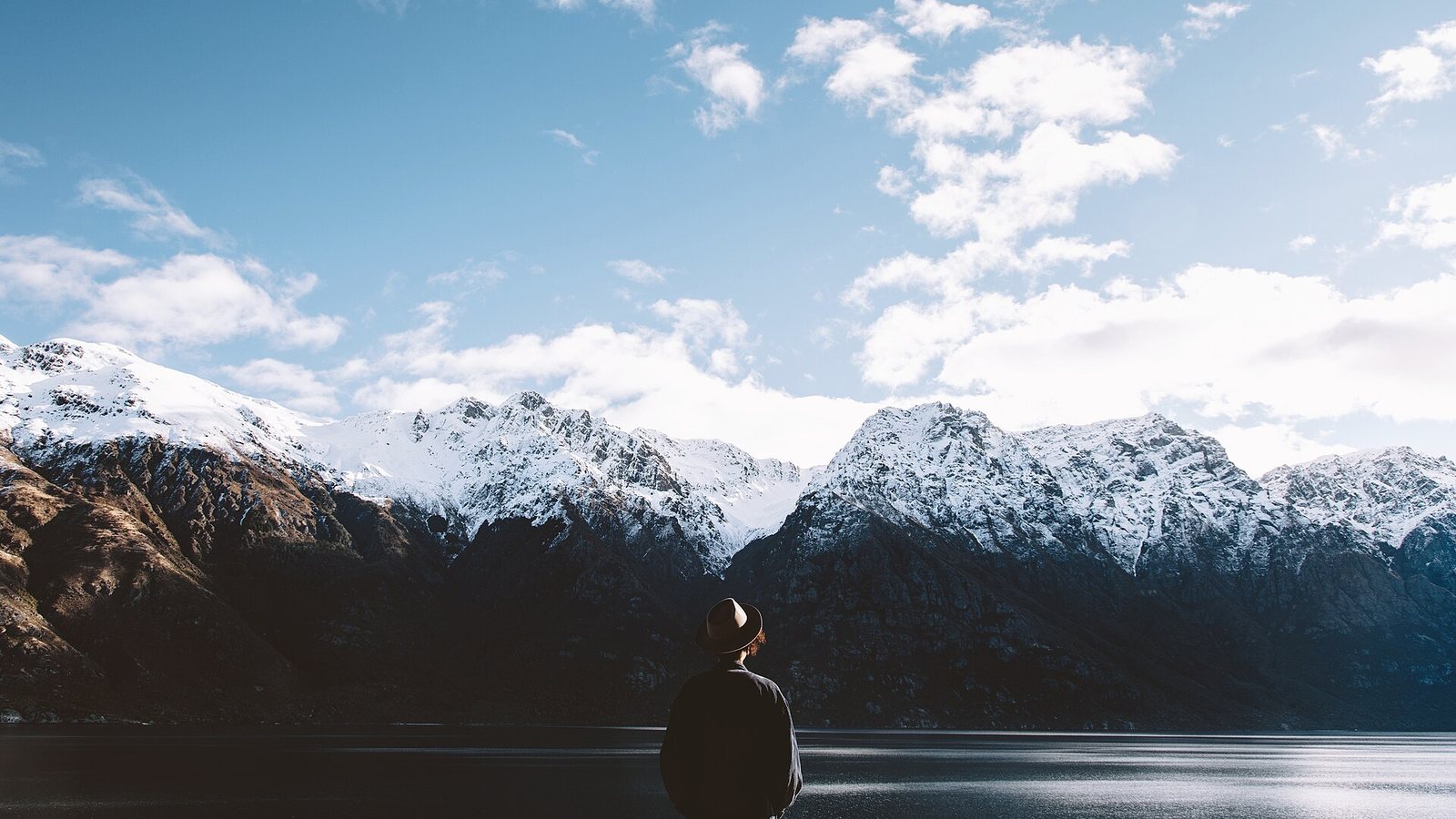 A person in a hat stands with their back to the camera, looking across a dark lake towards snow-covered mountains under a blue sky with clouds.
