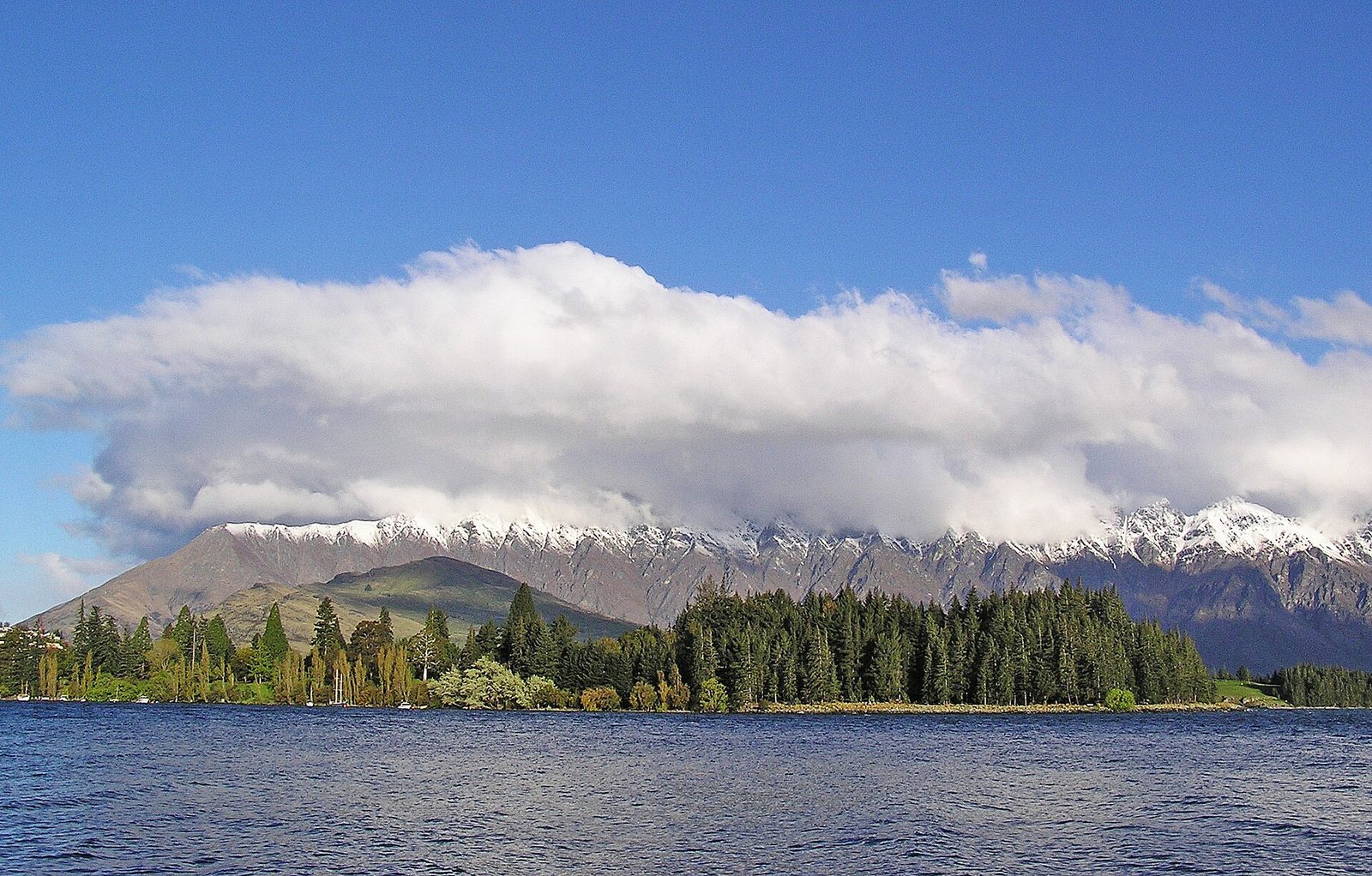 A wide view across a lake to a tree-lined shore, with snow-capped mountains partially covered by clouds under a blue sky in the background.