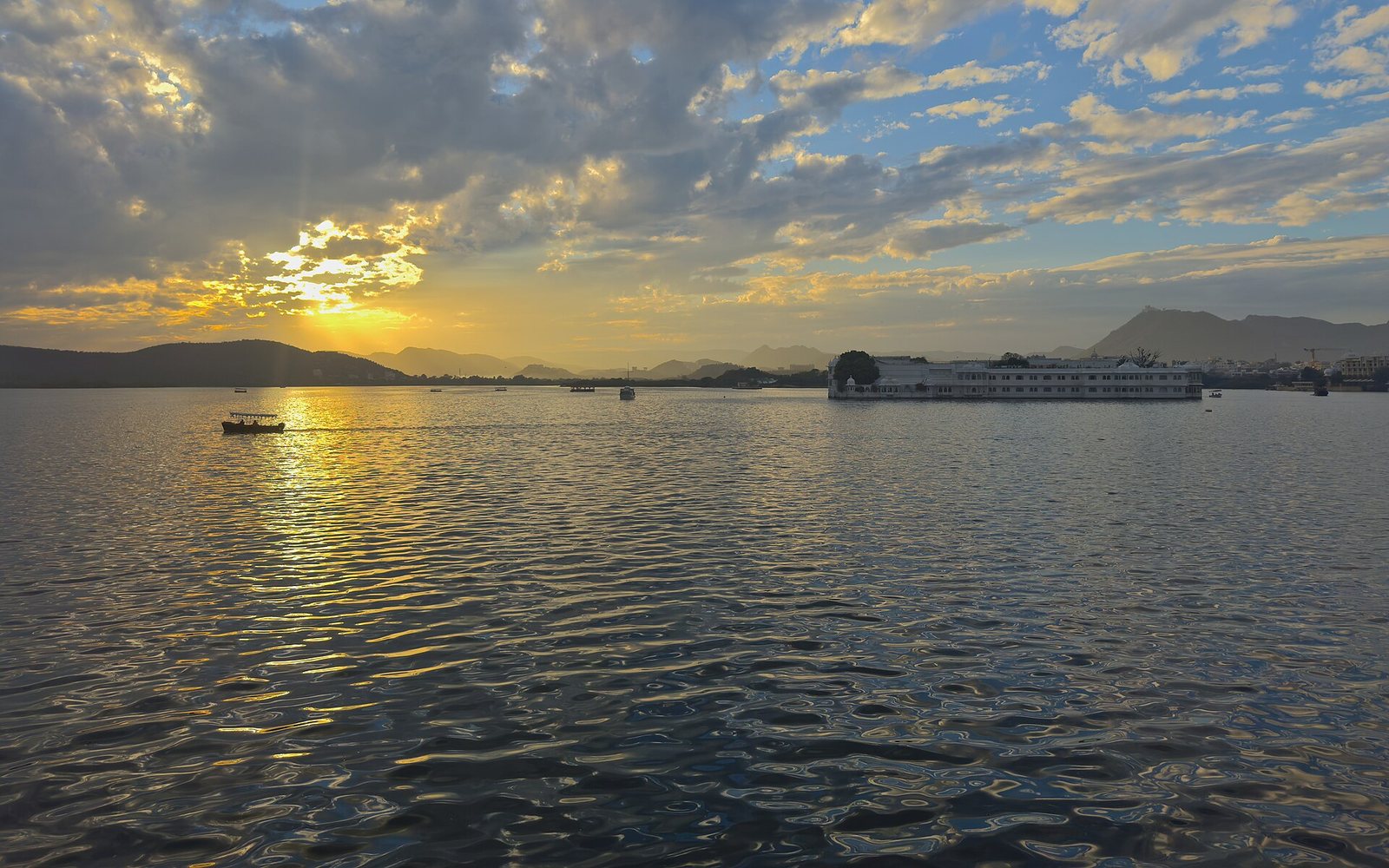 A sunset view over Lake Pichola, showing a boat on the left and the Lake Palace building on the right, with hills in the distance.