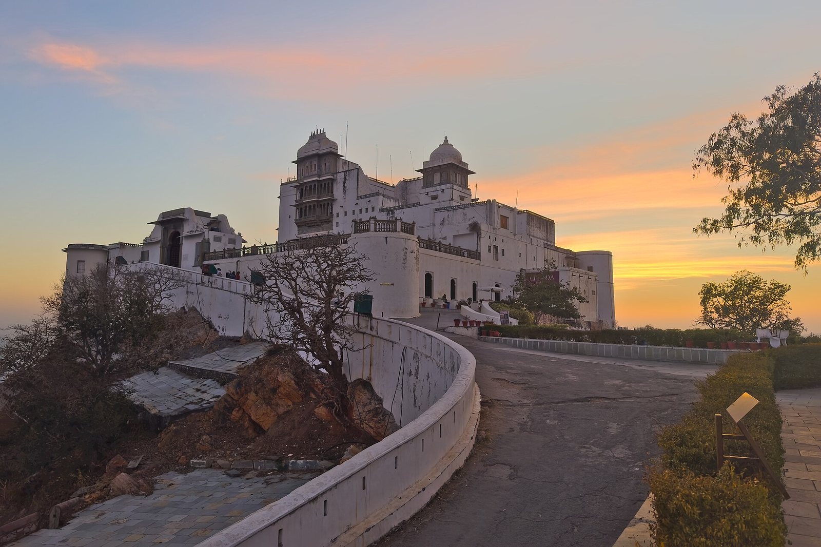A white palace structure with domes and balconies stands on a hill, accessed by a winding road, against a sky with orange, pink, and blue hues at dusk.