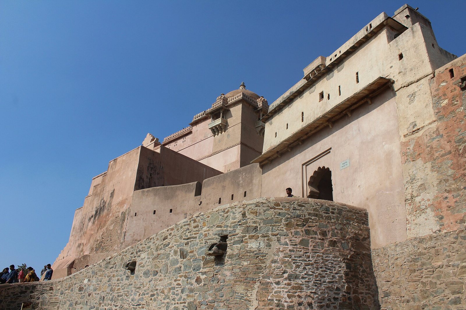 A large fort structure with stone and plastered walls rises against a clear blue sky, featuring a domed tower and an arched entrance.