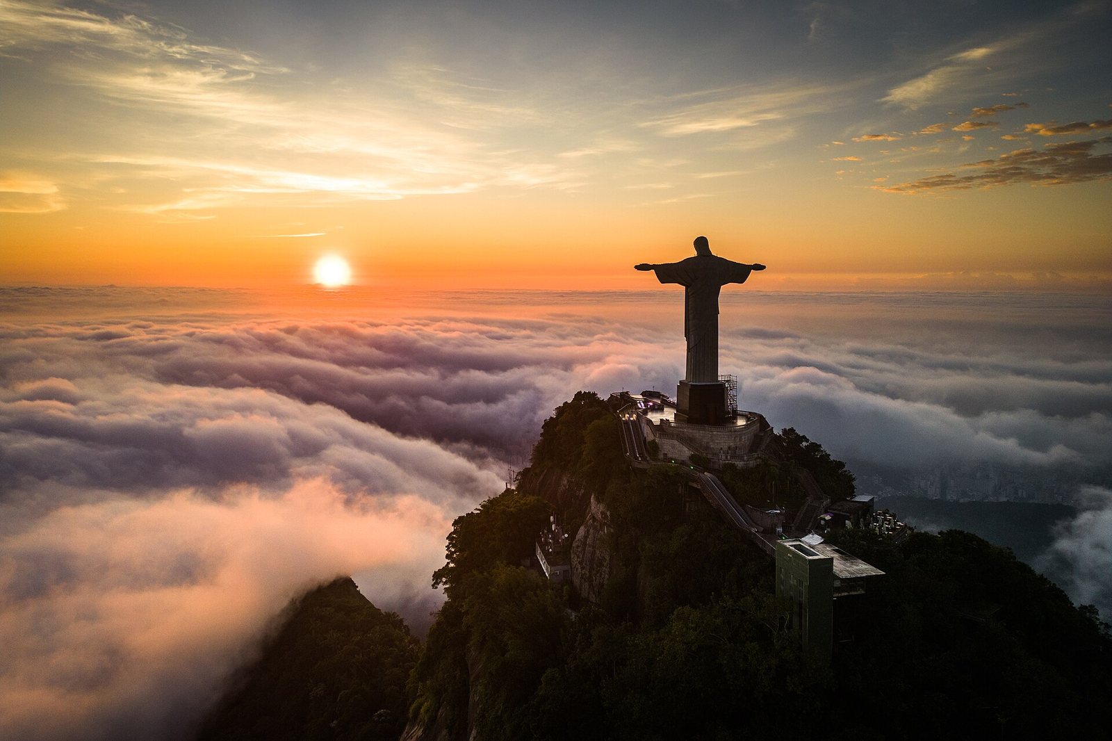 The Christ the Redeemer statue stands on a mountain peak, silhouetted against a sunset with clouds covering the landscape below.