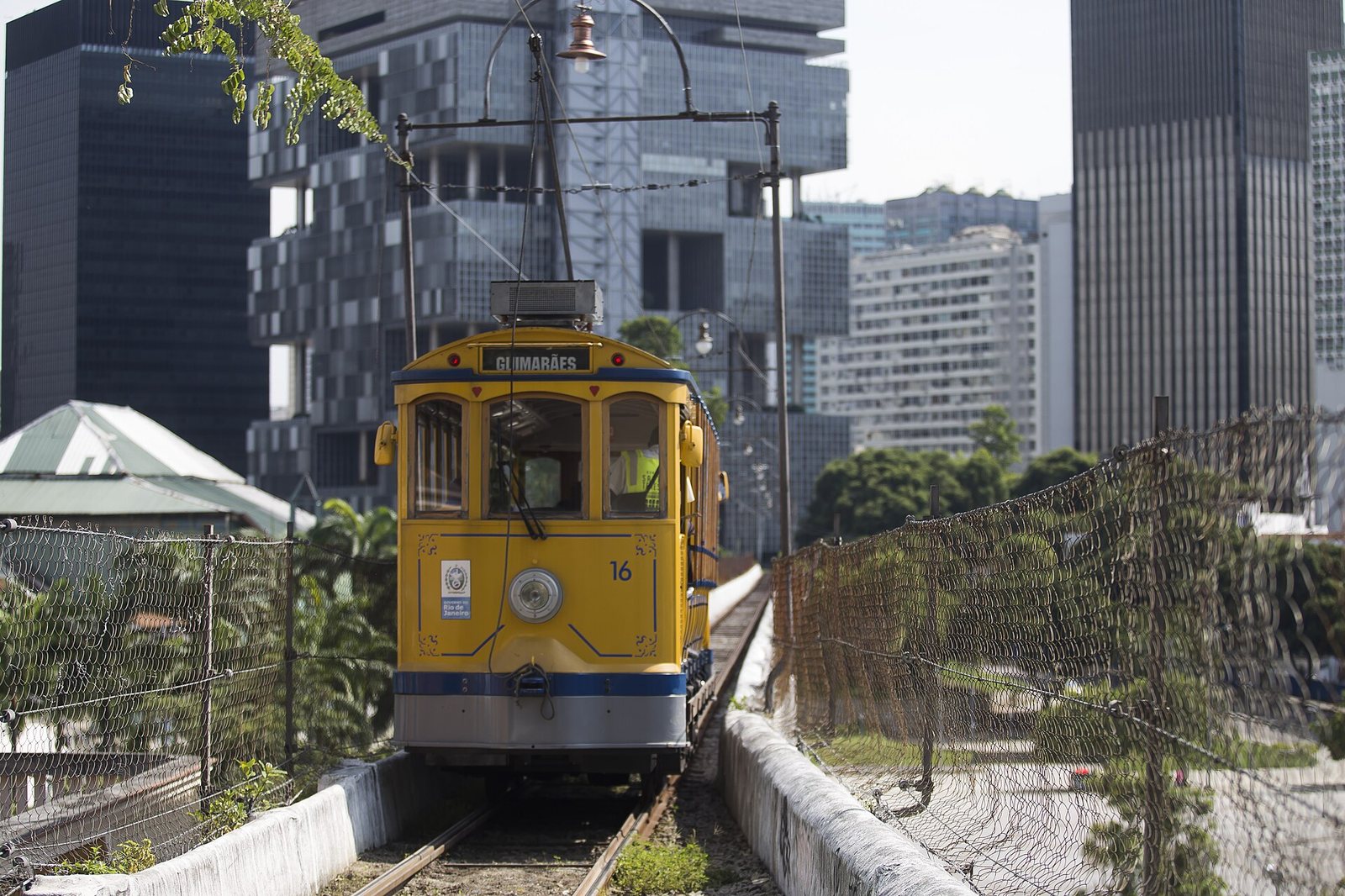 A yellow Santa Teresa tram on its track in Rio de Janeiro, with office towers of the centre behind.