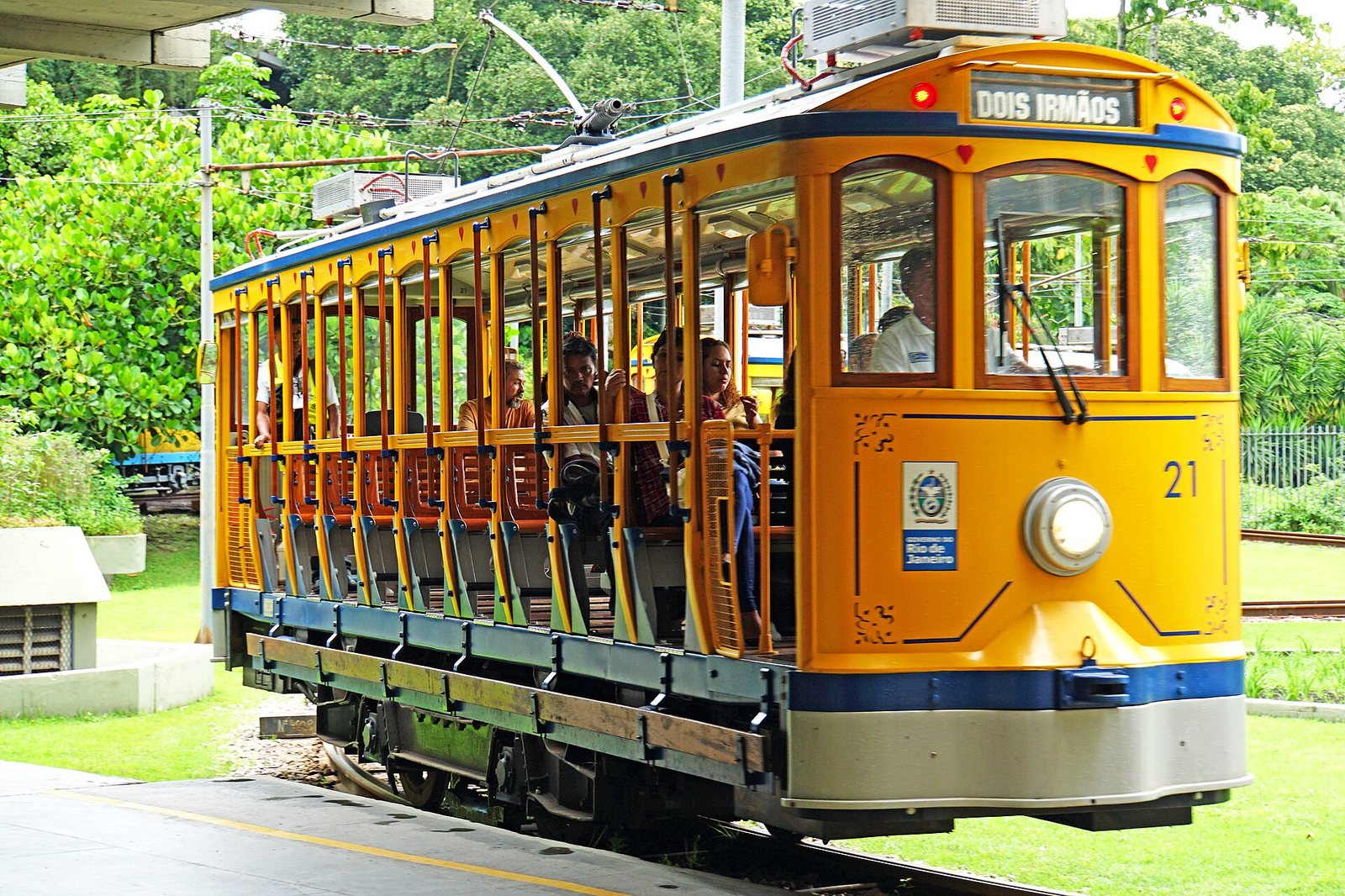 A yellow and blue tram, numbered 21 and displaying 'DOIS IRMÃOS', carries passengers on tracks with green foliage in the background.