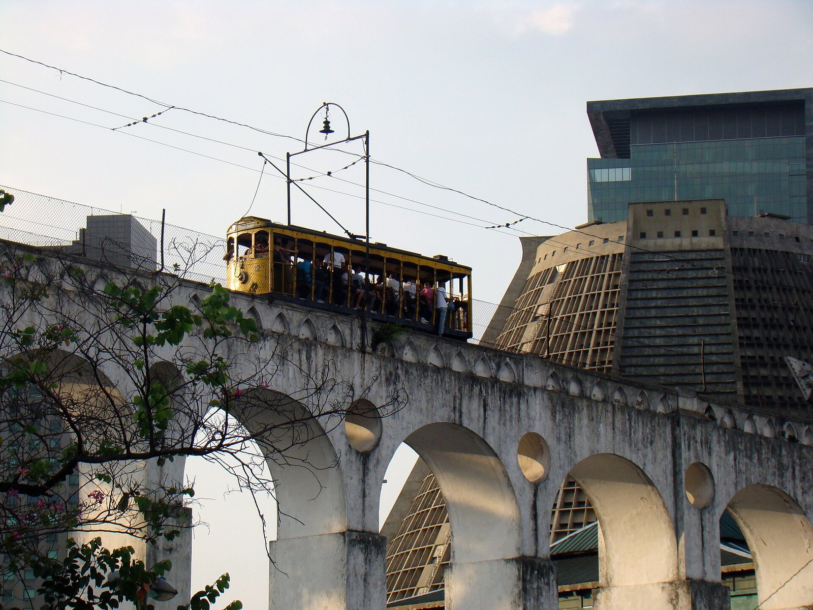 A yellow tram with passengers crosses a white arched aqueduct, with a large conical building and a modern glass building in the background.