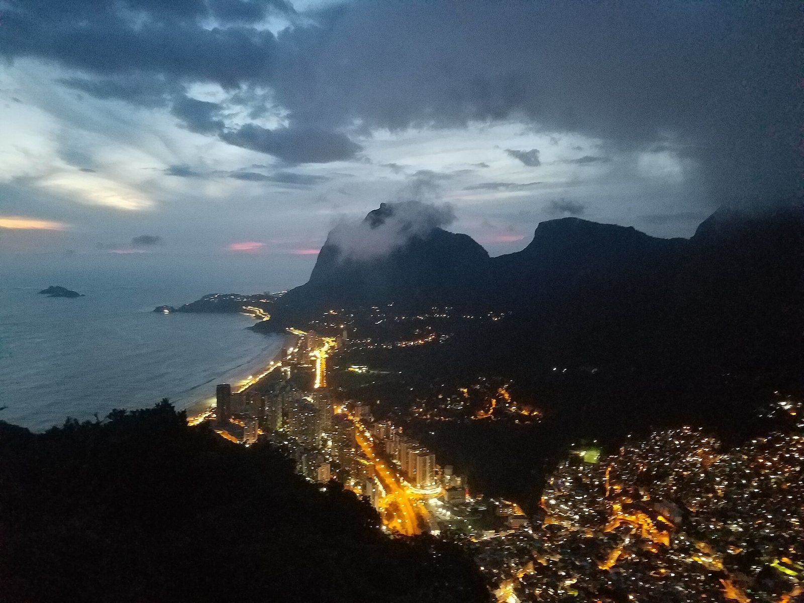 An aerial view of a coastal city at dusk, showing illuminated buildings along a beach, dark mountains with clouds, and an island in the sea under a cloudy sky.
