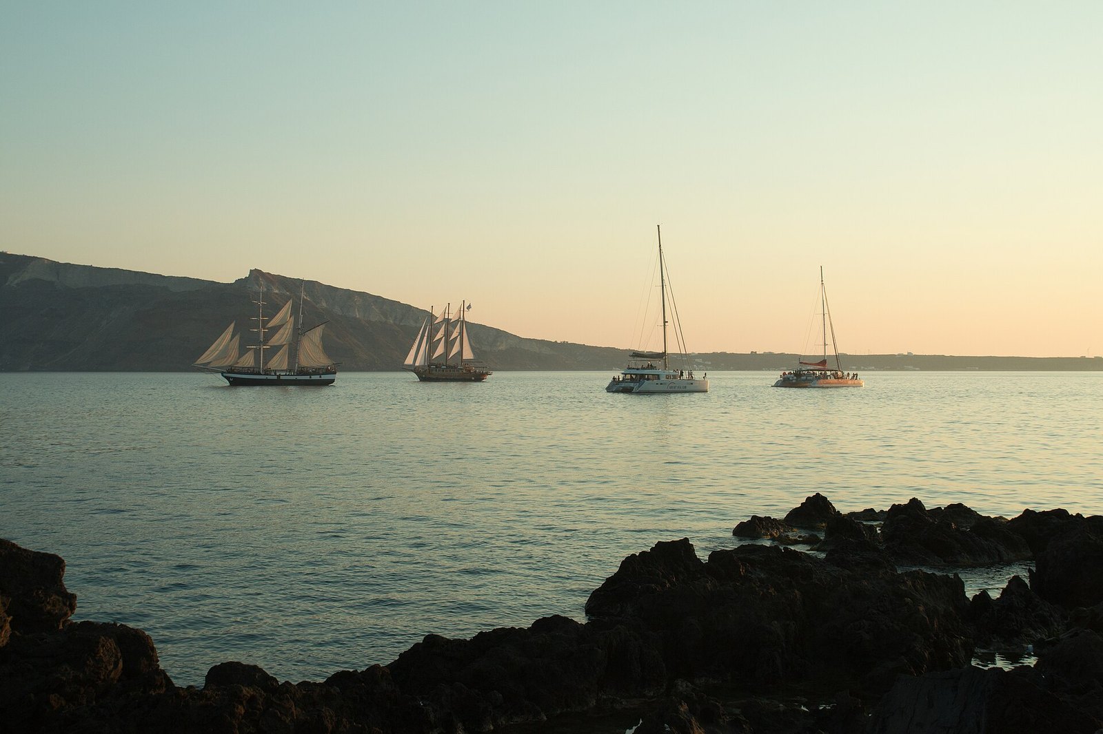 Three tall ships and a catamaran at anchor on calm water at sunset, with dark rocks in the foreground and a headland beyond