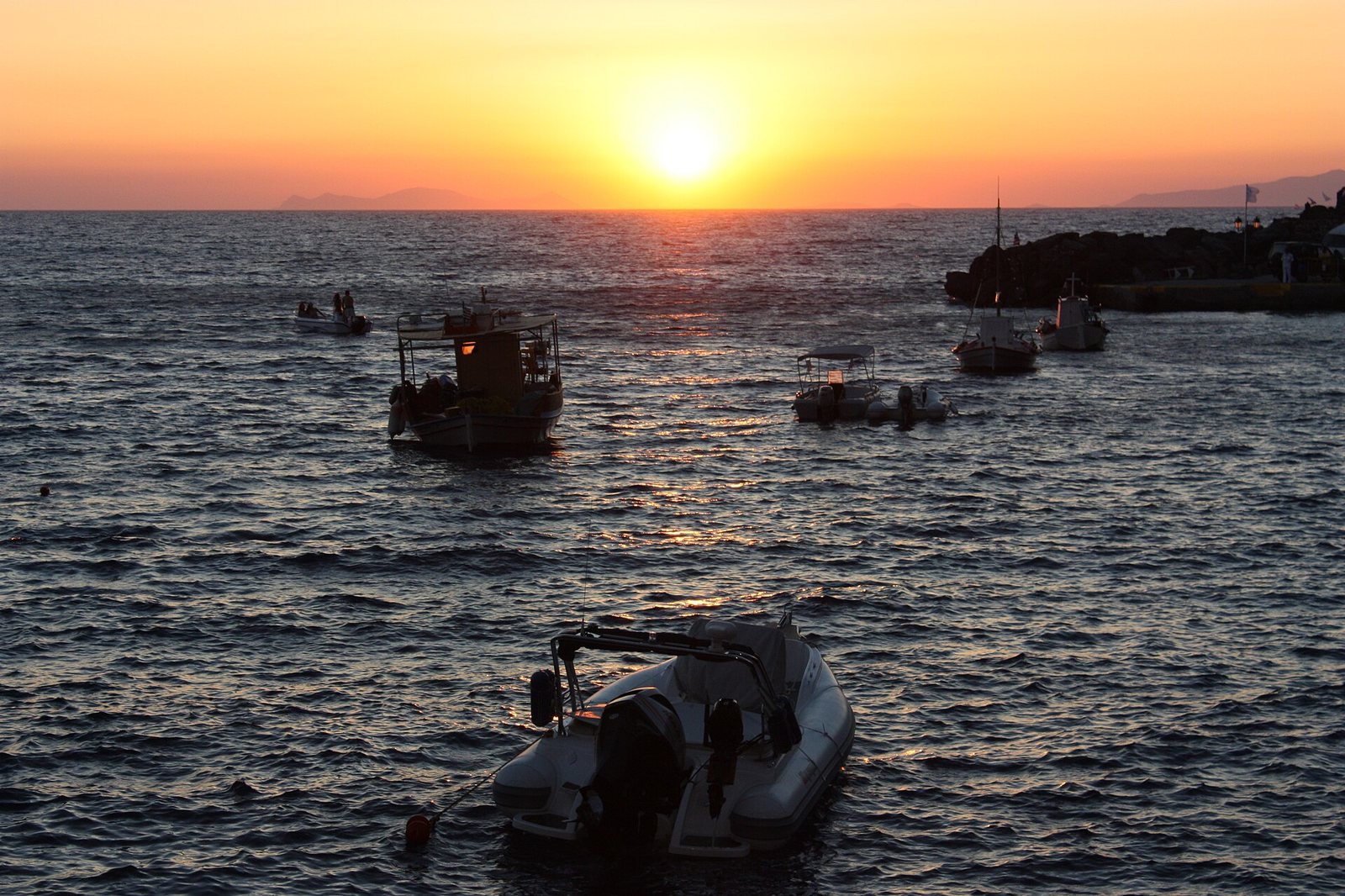 A sunset over water with several boats moored in the foreground and midground, and distant land visible on the horizon.