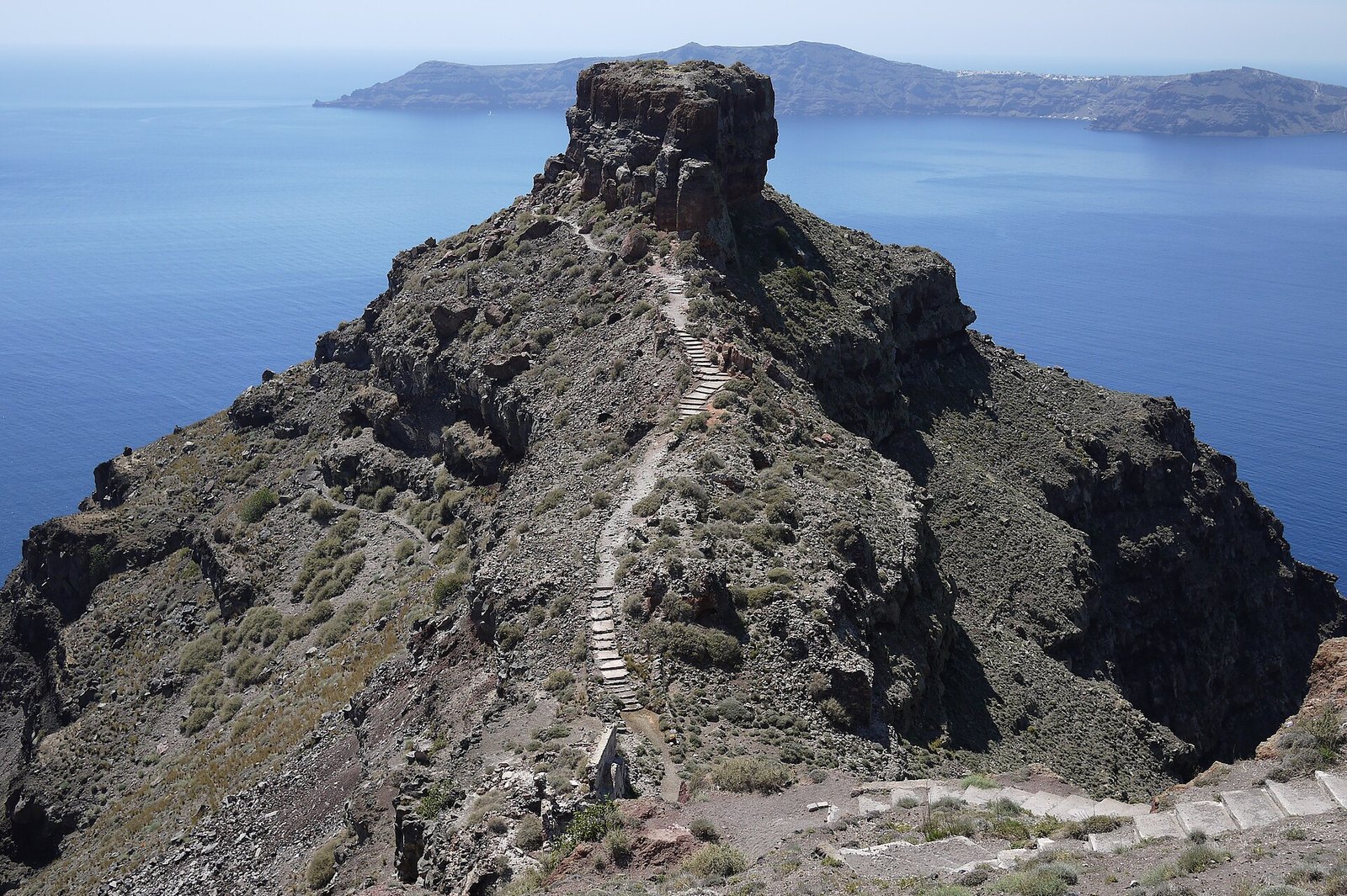 A steep rock outcrop with a stepped path along its spine rising above the blue sea of the caldera, with a distant island on the horizon