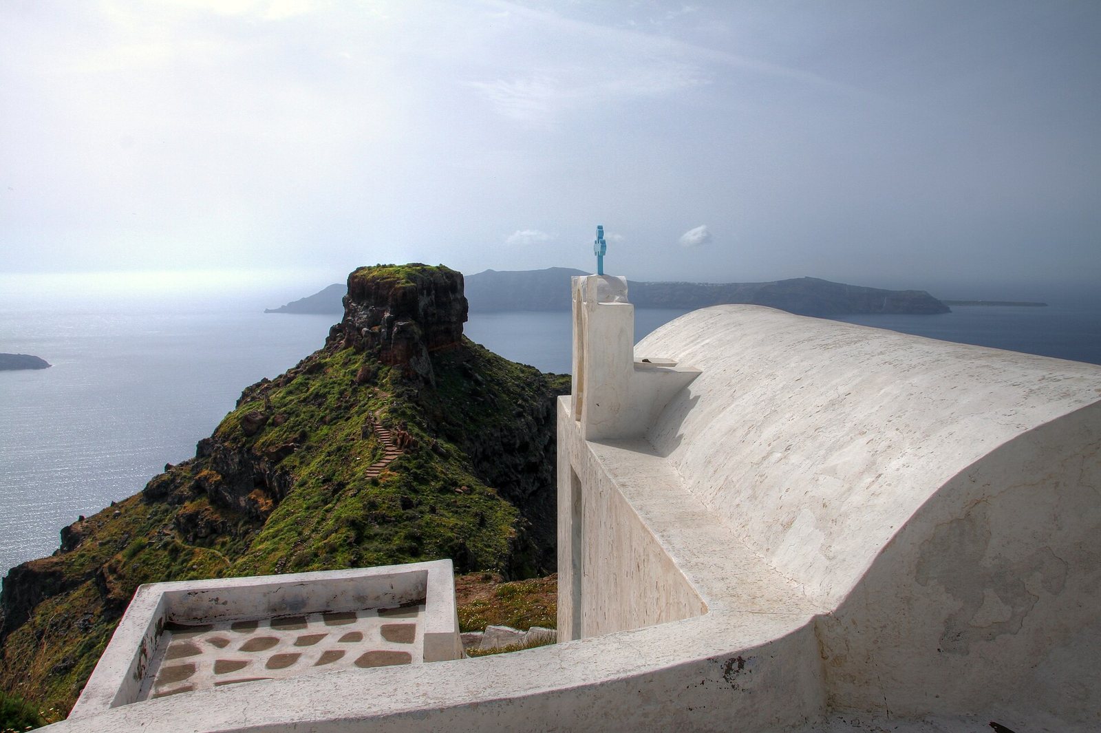 A white church with a blue cross on its roof stands in the foreground, overlooking a green, rocky outcrop and the sea with distant landmasses under an overcast sky.