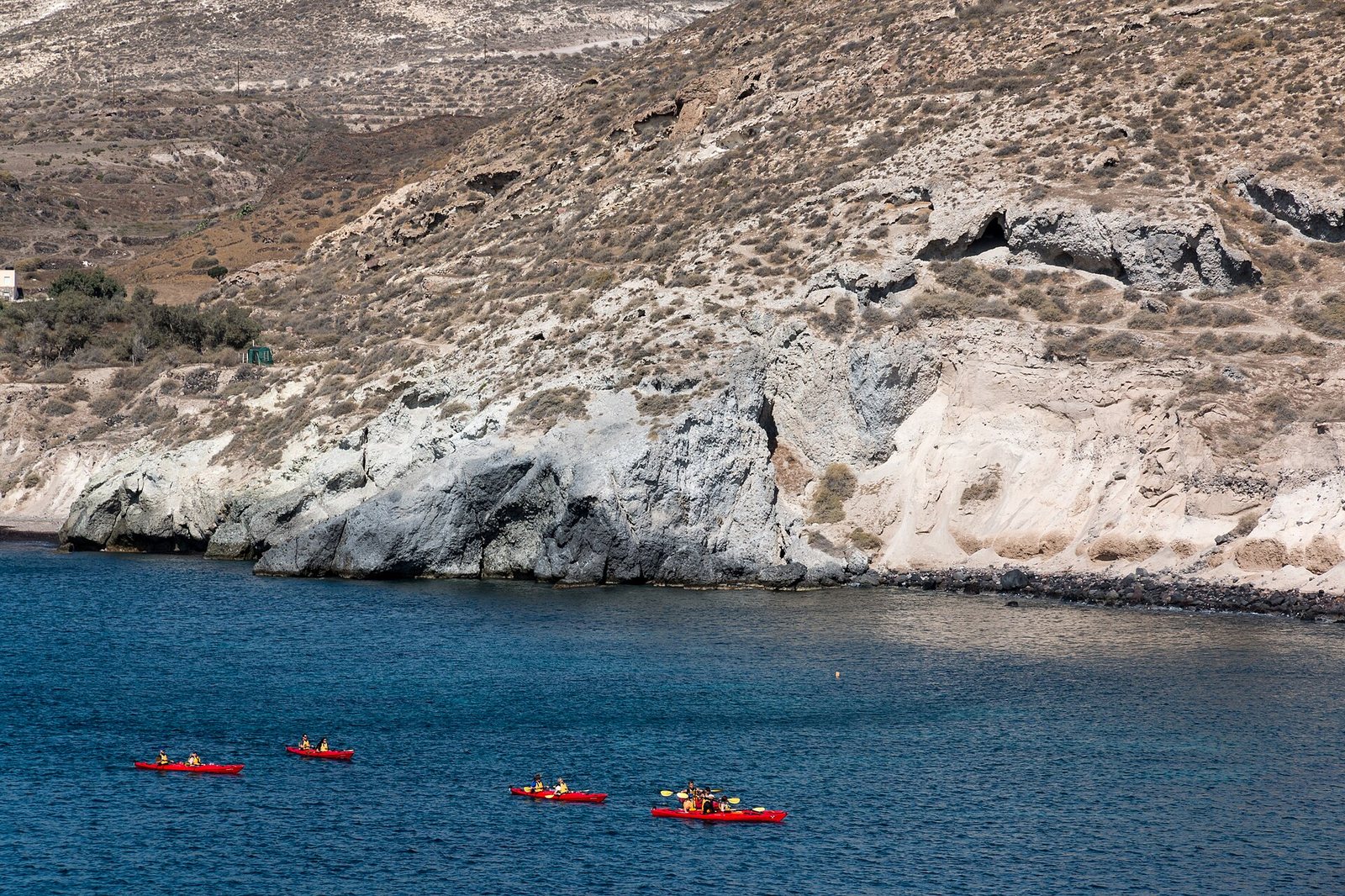 Four red kayaks with people paddle on blue water near a rocky coastline with dry, scrub-covered hills rising behind it.