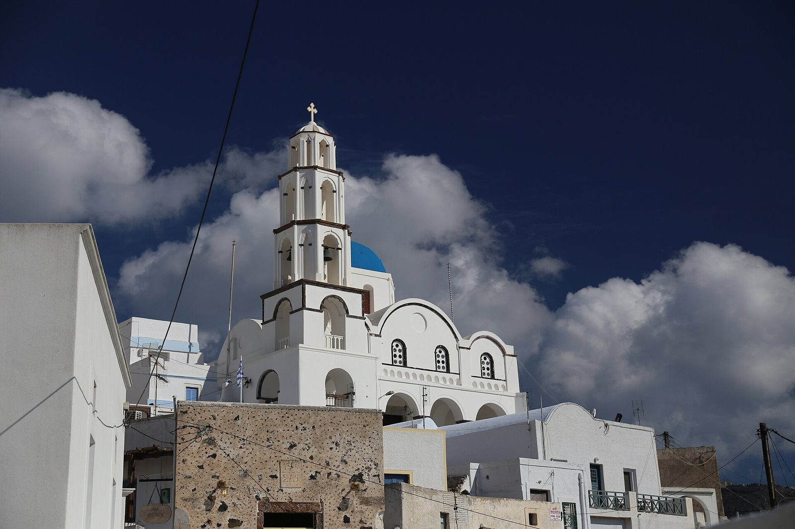 A white church with a blue dome and a tall bell tower stands against a dark blue sky with white clouds, surrounded by other white buildings and a stone wall.