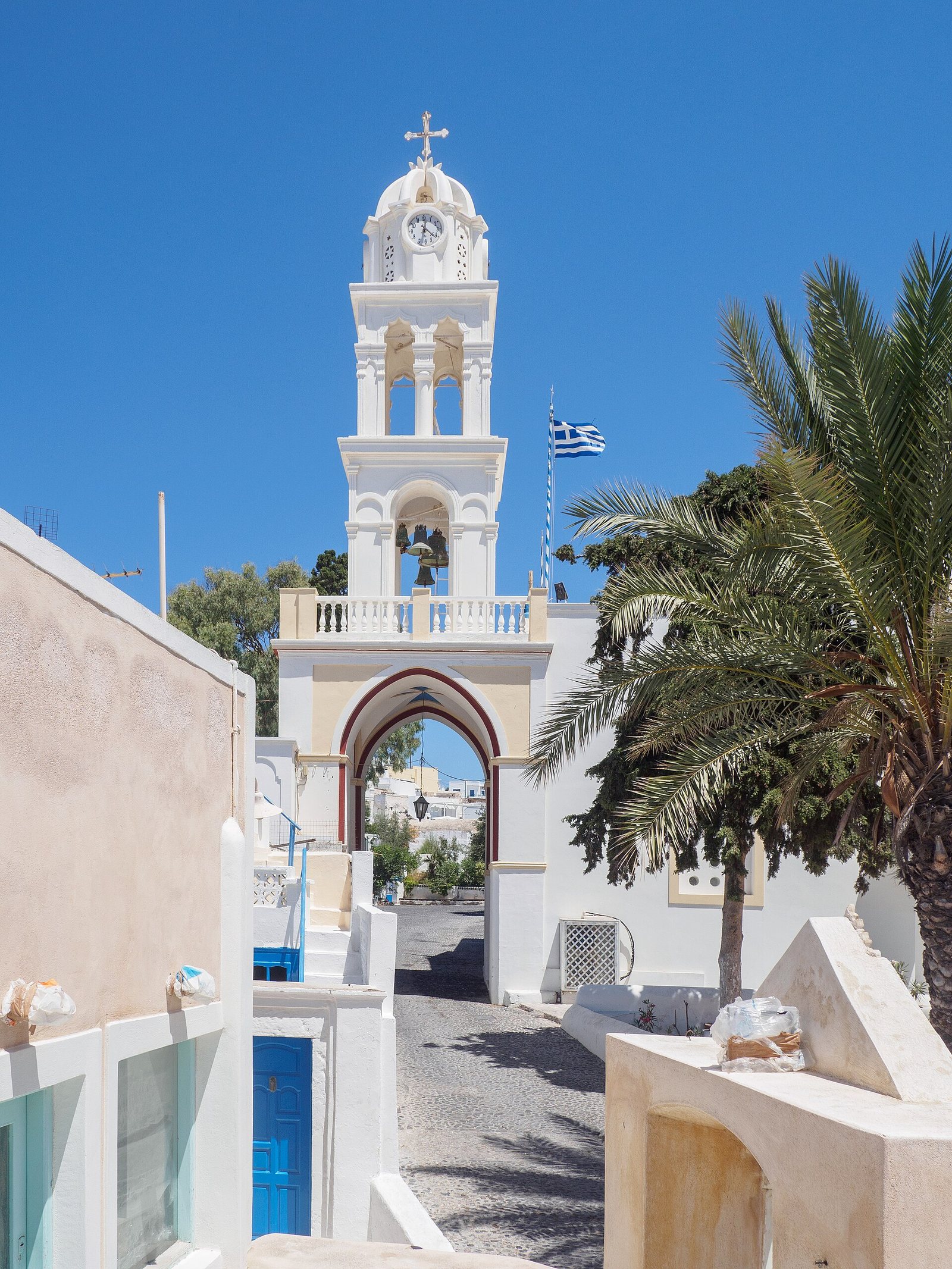 A white bell tower with a clock and cross stands over an arched gateway, flanked by white buildings, a blue door, and palm trees under a clear sky.