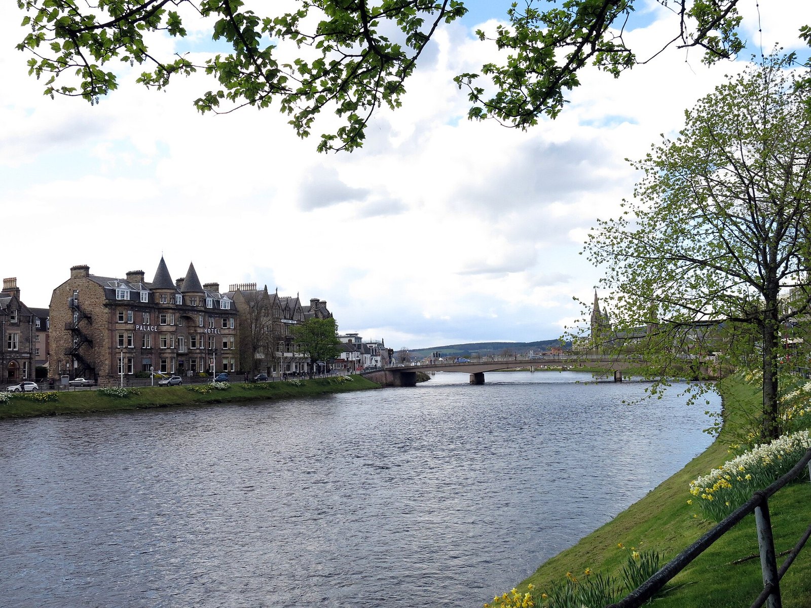 A river flowing past a row of stone buildings, including a hotel with two turrets, with a bridge beyond and daffodils on the bank
