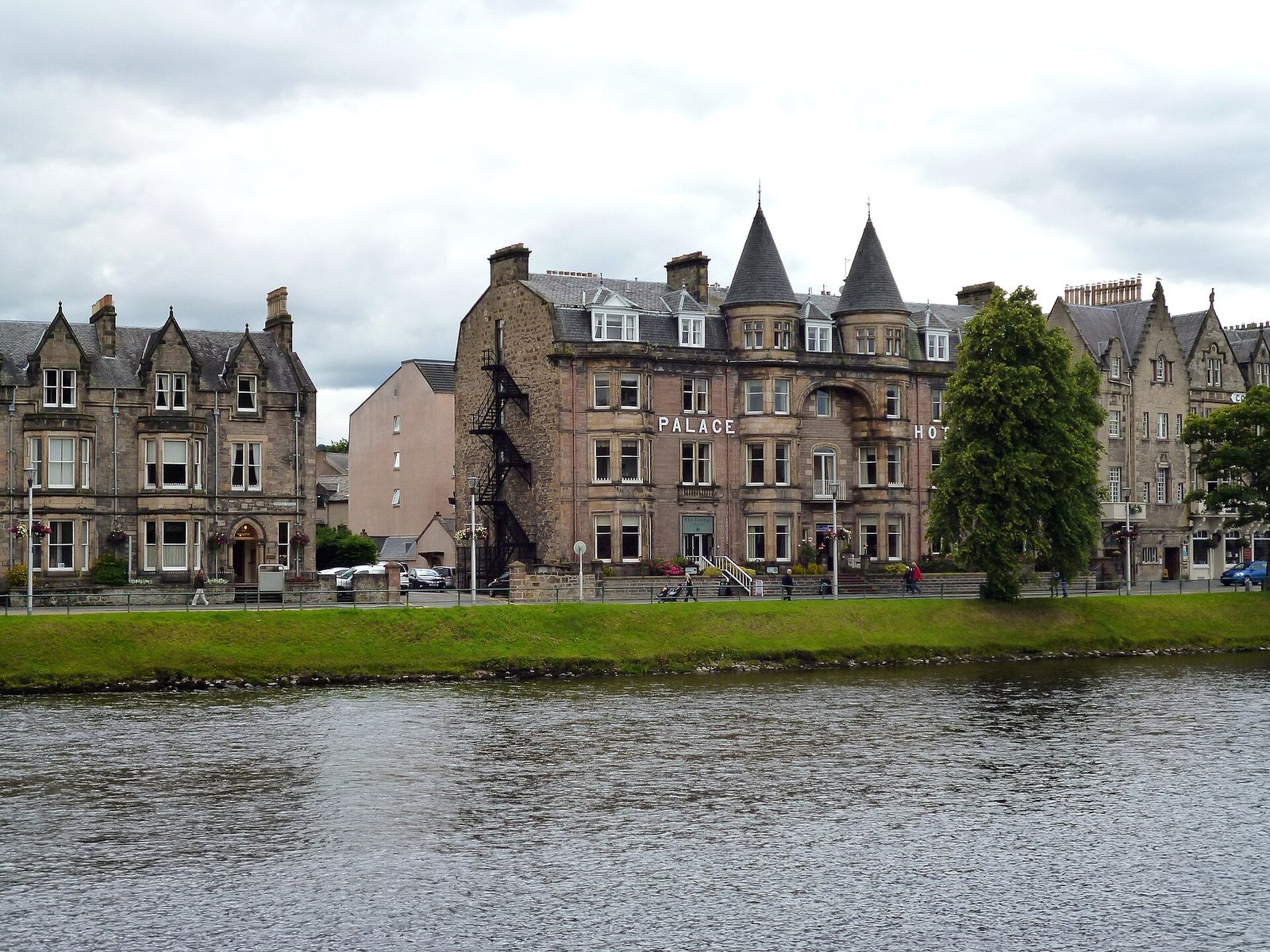 A river flows in the foreground, with a grassy bank leading to a row of stone buildings, including the Palace Hotel with two turrets, under an overcast sky.