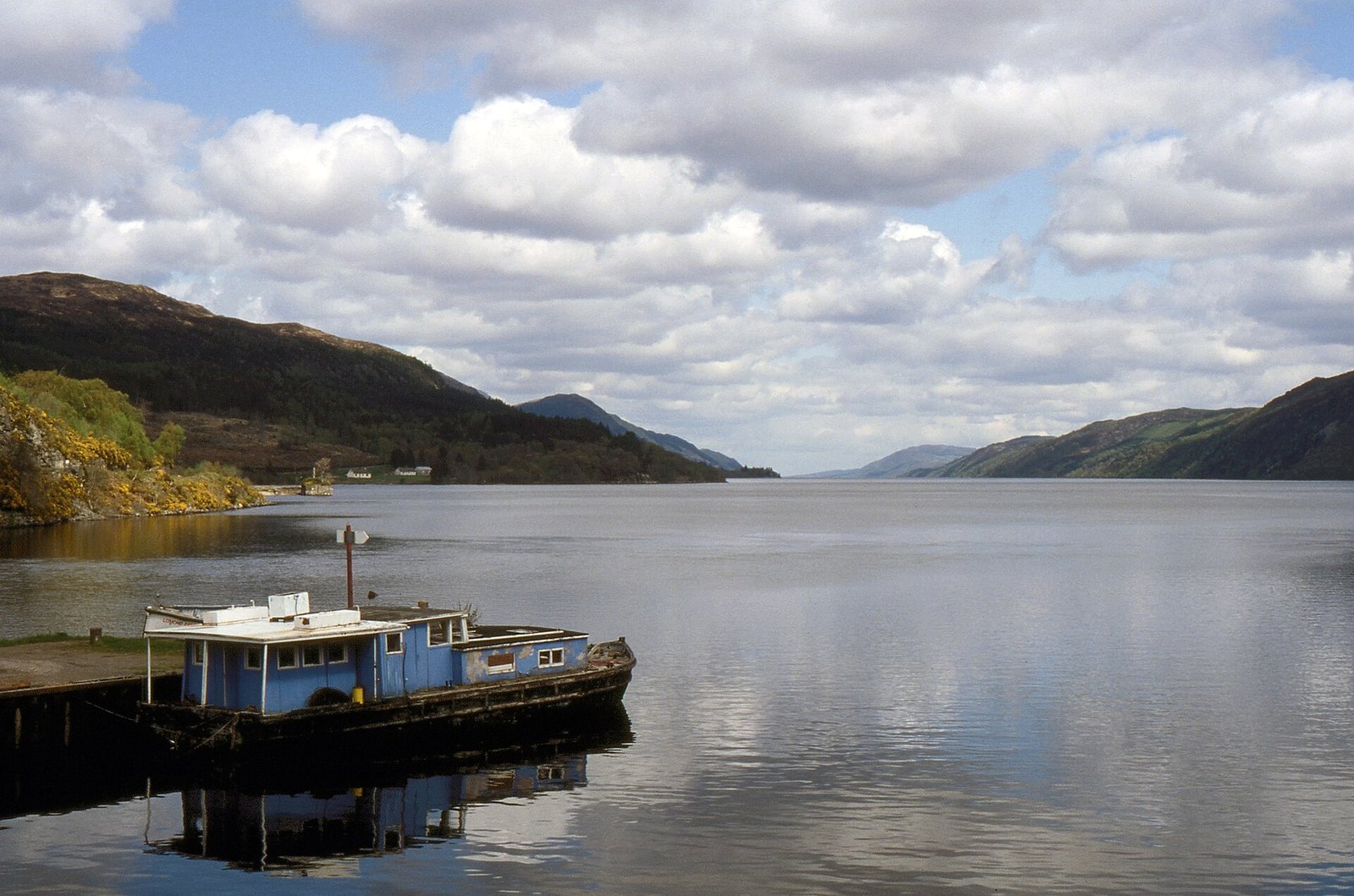 A grey-blue loch with a small boat moored at a jetty and hills on both shores under a cloudy sky