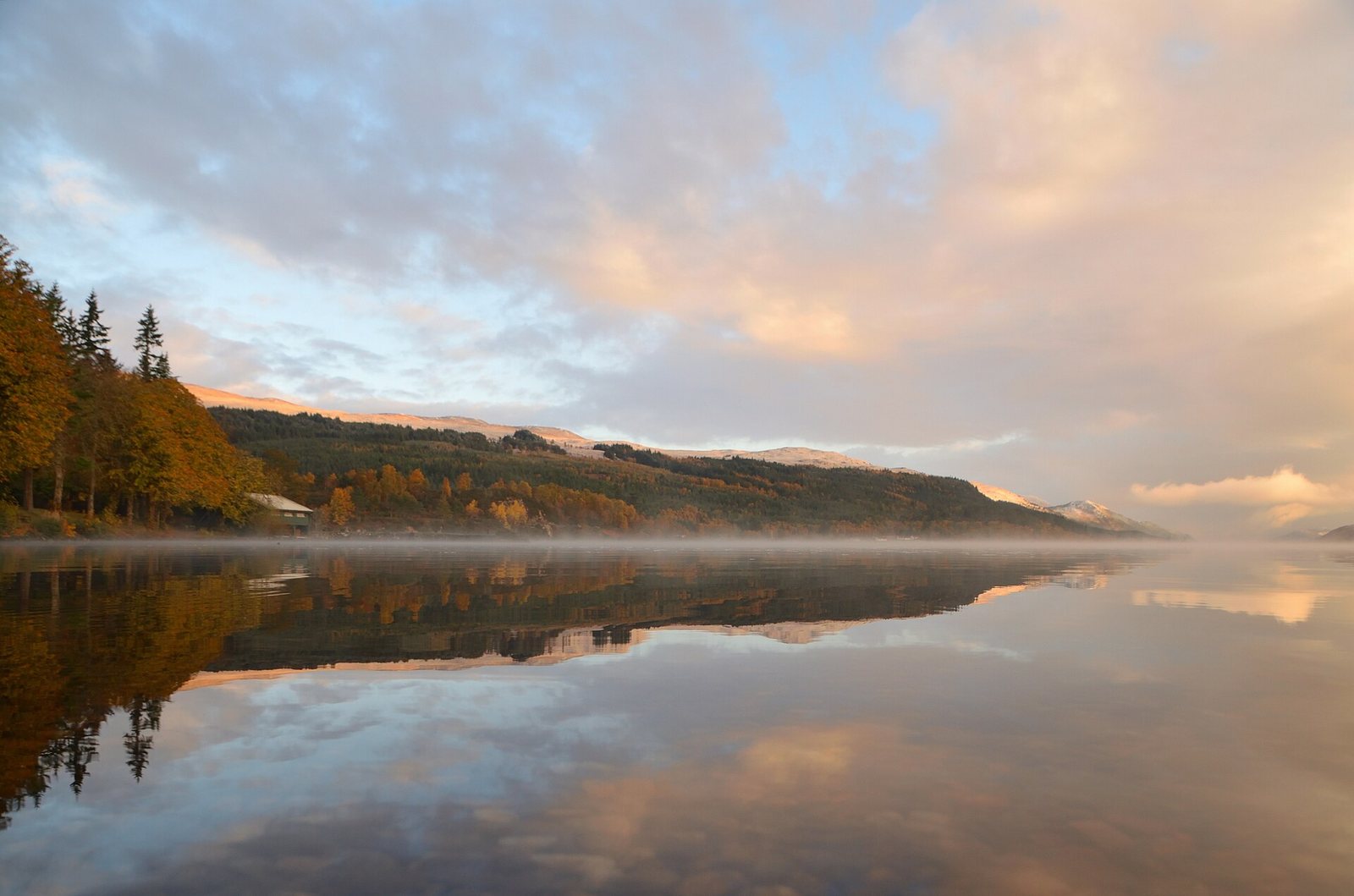 A loch with mist reflecting a cloudy sky and treelined hills, with a small building visible on the left shore.