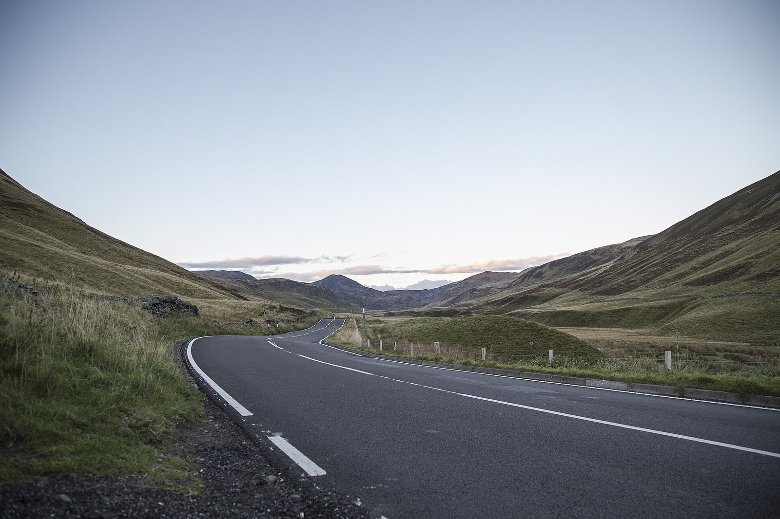 A winding tarmac road with white lines curves through a valley, flanked by grassy hills, under a pale sky with distant mountains.