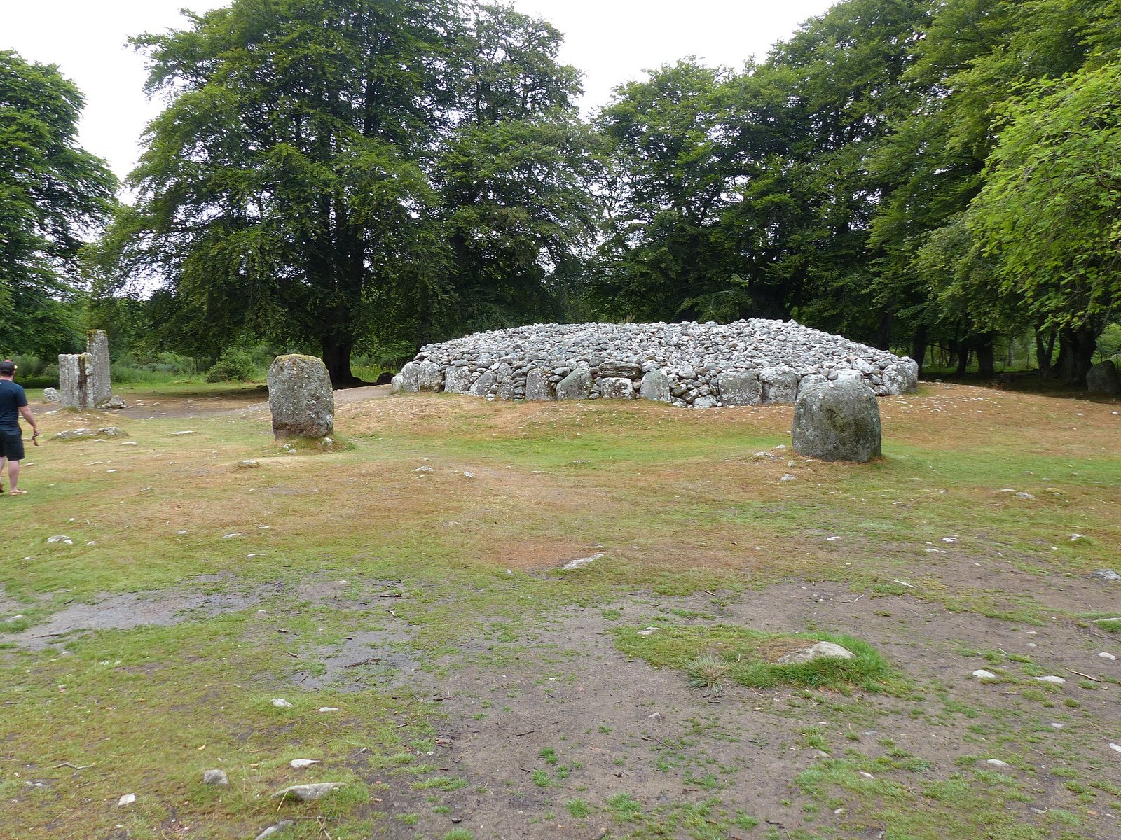 A large stone cairn between standing stones on grassy ground, with tall trees behind