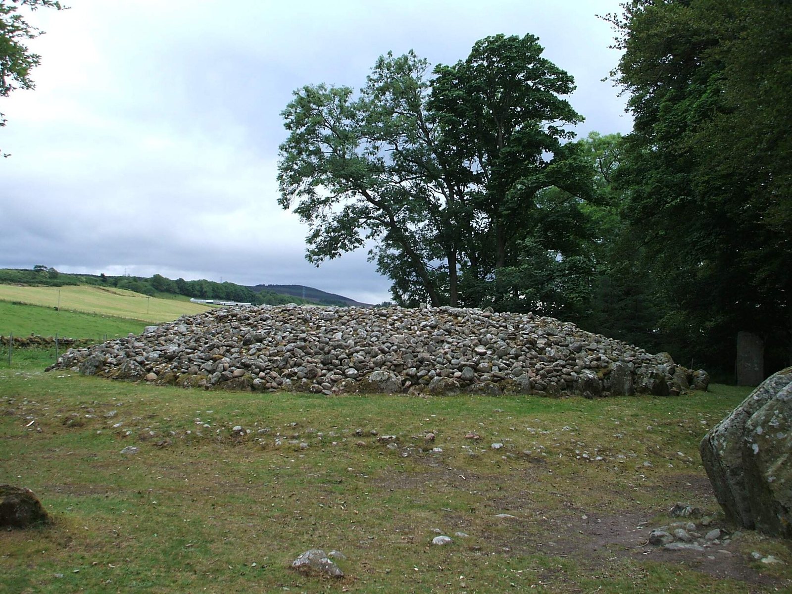 A large stone cairn and a standing stone are visible on grassy ground, with trees and distant green fields under an overcast sky.