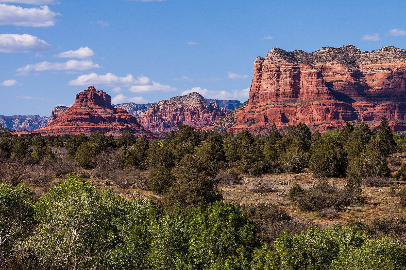 Bell Rock and other red sandstone formations rise above green trees and shrubs under a blue sky with white clouds.
