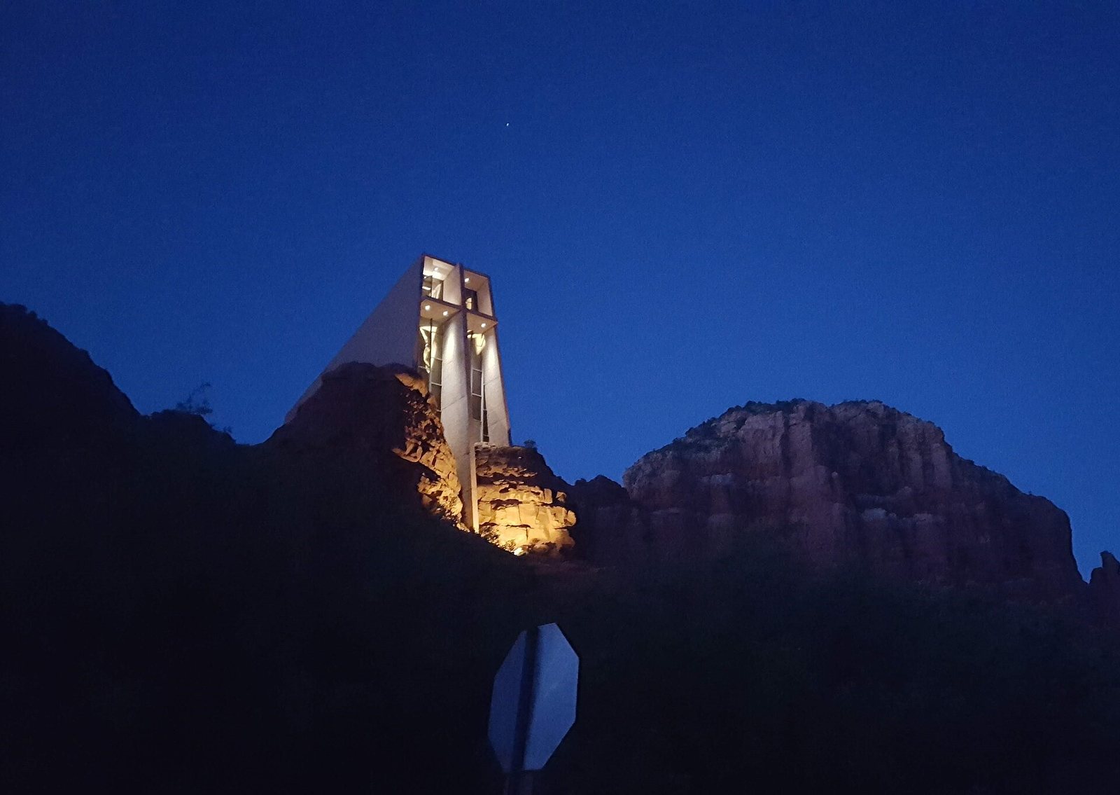 A modern chapel, illuminated at night, stands on a rocky outcrop against a dark blue sky. Other dark rock formations are visible in the background.