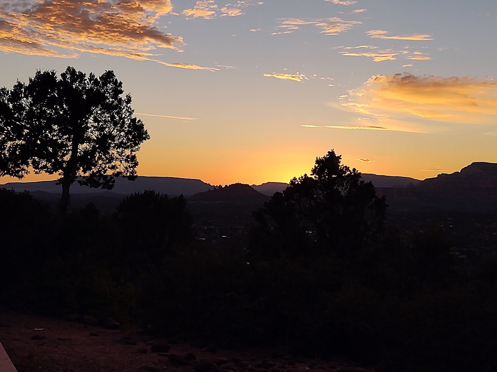 A sunset over a landscape with silhouetted trees in the foreground and distant hills under an orange and blue sky with clouds.