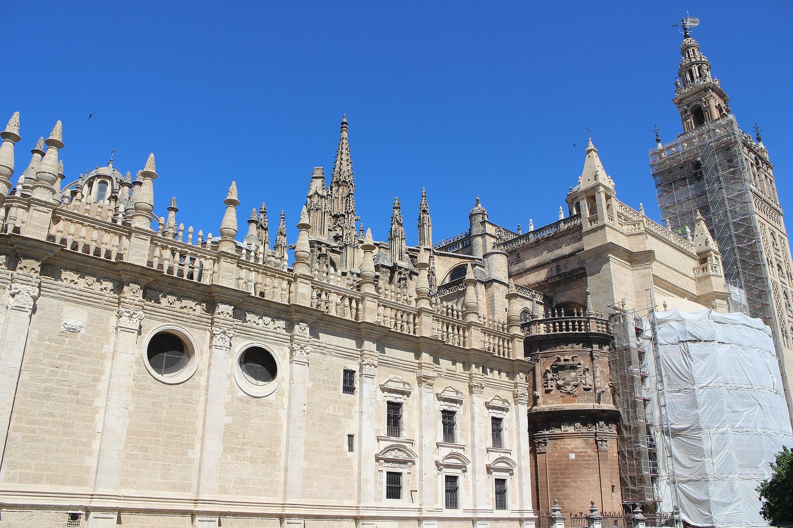 The exterior of a large, ornate stone cathedral with multiple spires and a tall tower, partially covered in scaffolding, under a clear blue sky.