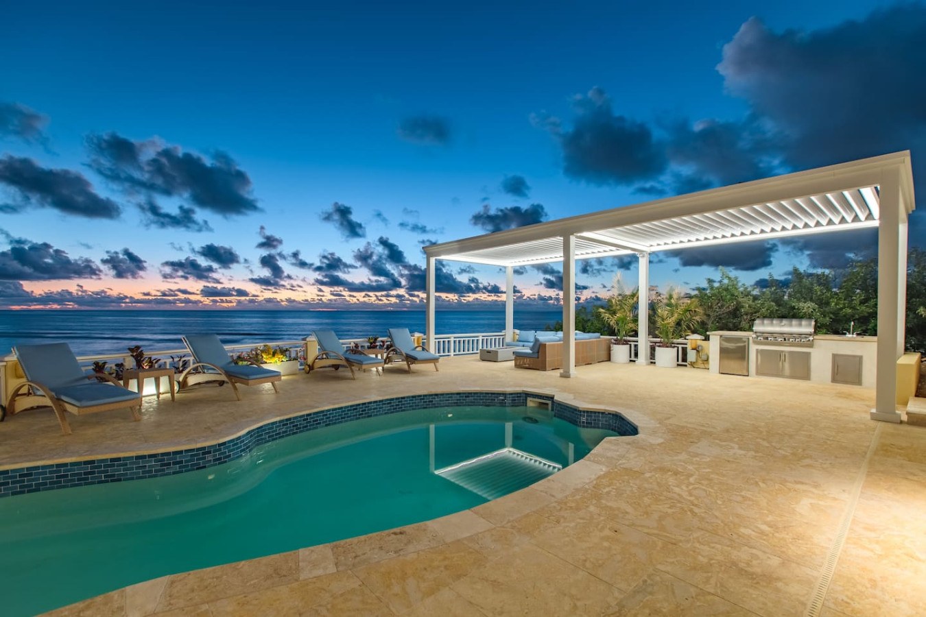 The pool deck of The Cerulean villa at twilight, with loungers, a white pergola and an outdoor kitchen above the Caribbean Sea.