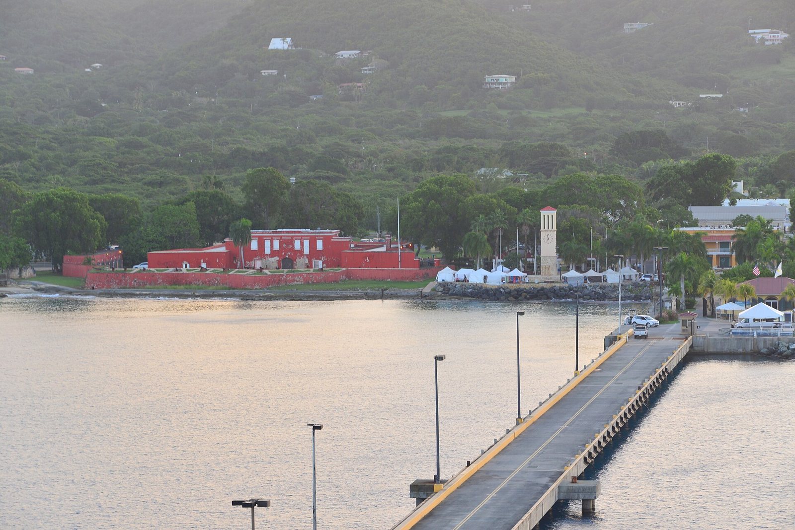 The red walls of Fort Frederik on the Frederiksted waterfront, with the long cruise pier in the foreground and green hills behind.