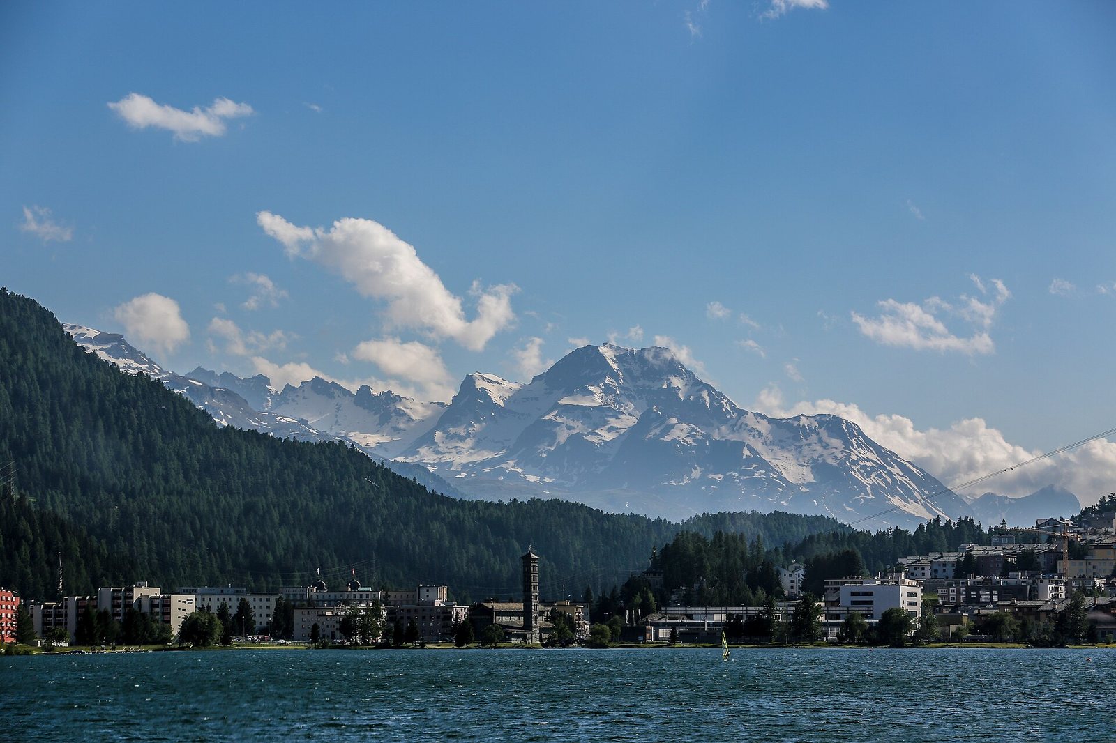 A lake with a sailboat in the foreground, a town with buildings and a tower along its shore, forested hills, and snow-capped mountains under a blue sky.