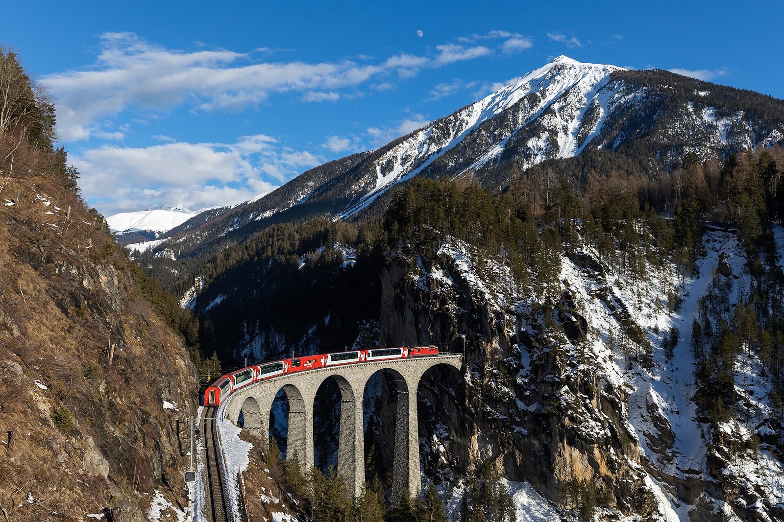 A red and white Glacier Express train crosses the Landwasser Viaduct, surrounded by snow-covered mountains under a blue sky with a crescent moon.