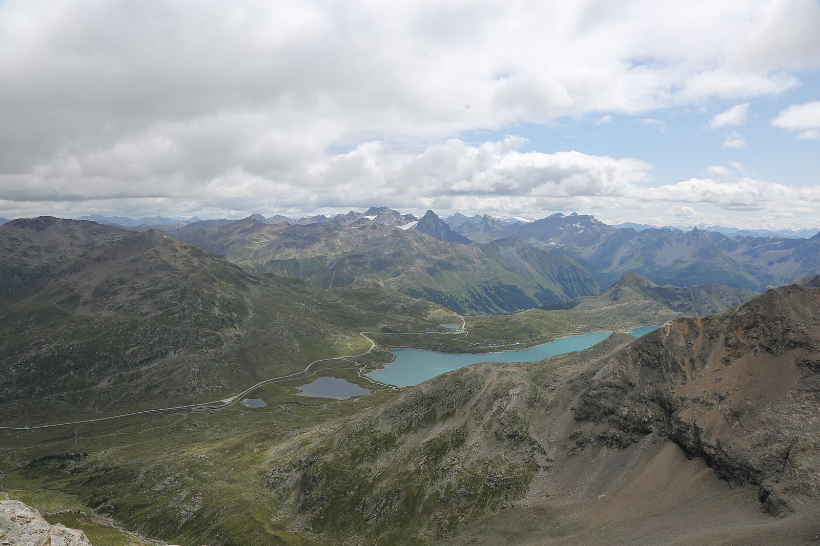 A panoramic view of multiple mountain ranges under a cloudy sky, with a large light blue lake (Lago Bianco) and smaller dark lakes, and a road winding through a green and rocky valley.