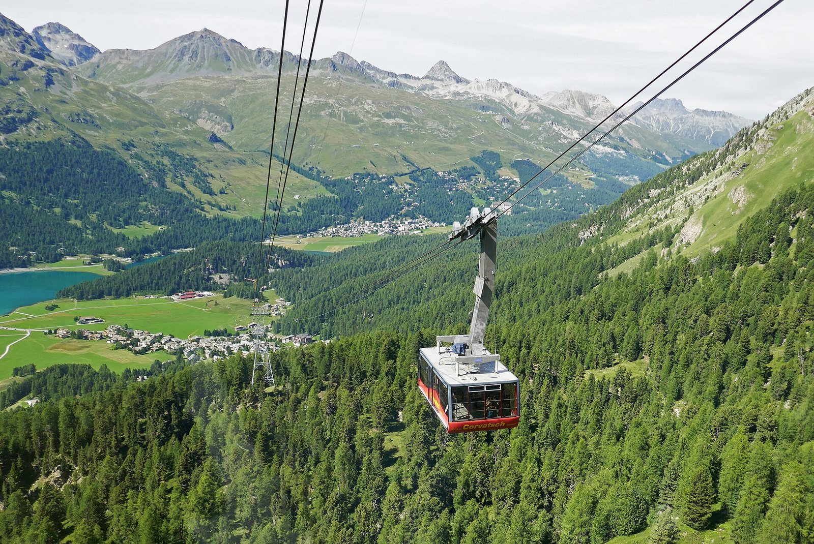 A red and yellow Corvatsch cable car cabin travels over a dense green forest, with a valley, village, turquoise lake, and mountains in the background.