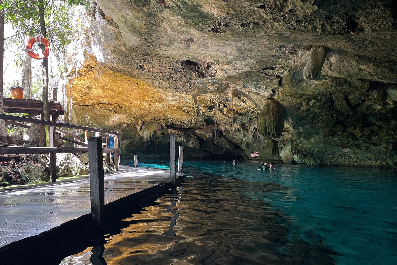A wooden walkway extends into a cave with clear blue water, where several people are swimming. Sunlight illuminates the cave entrance and rock formations on the ceiling.