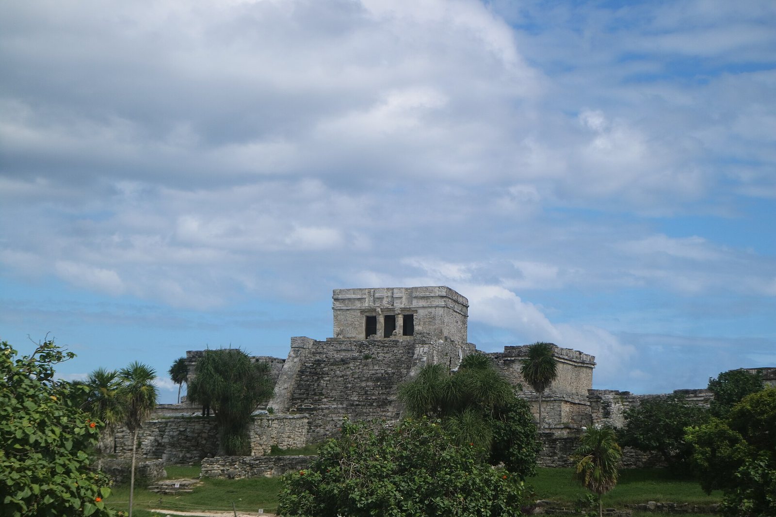 A stone Maya ruin, featuring a stepped pyramid structure topped with a building, is visible amidst green foliage under a partly cloudy sky.