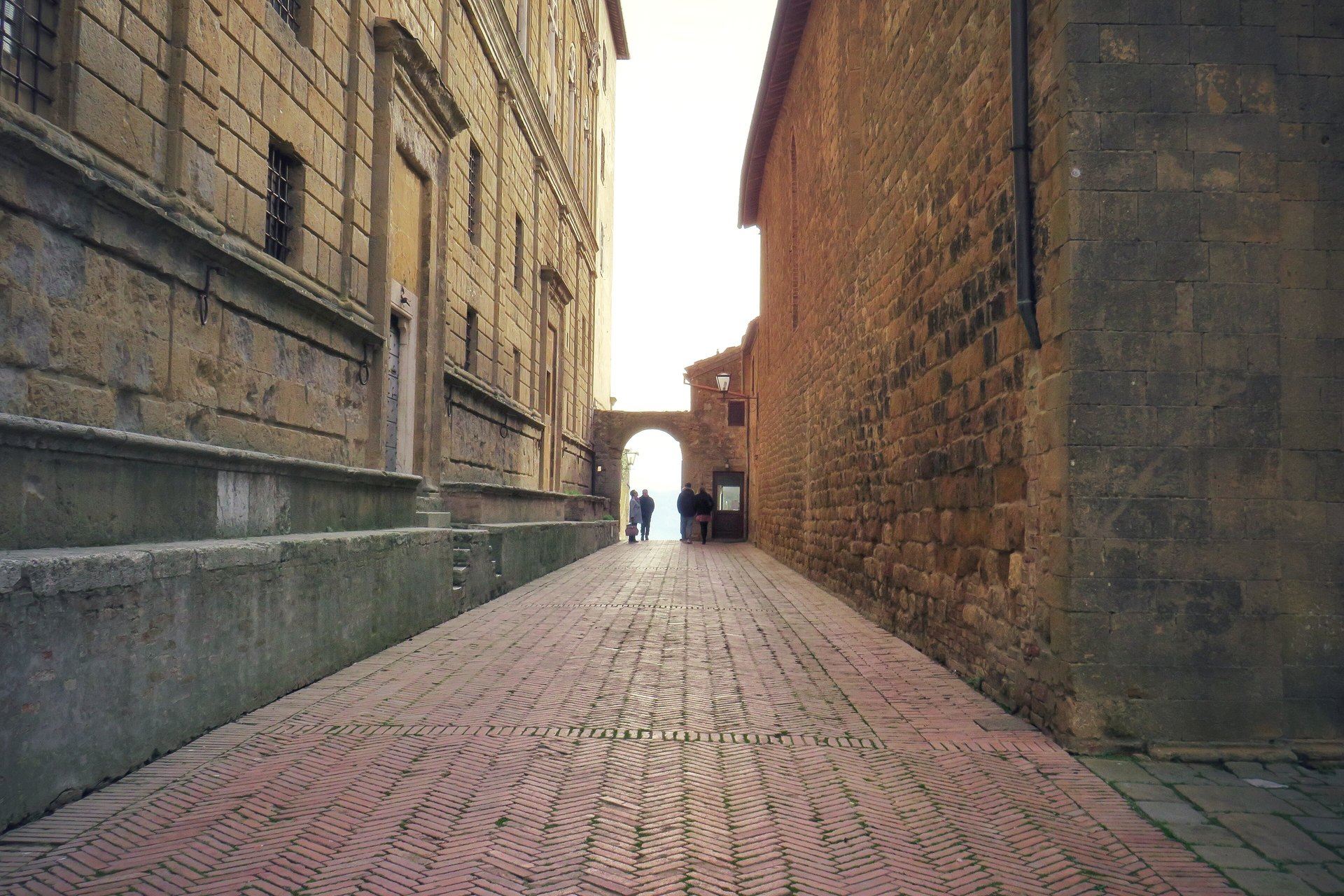 A narrow brick-paved lane between stone walls leading to an arch with a view beyond