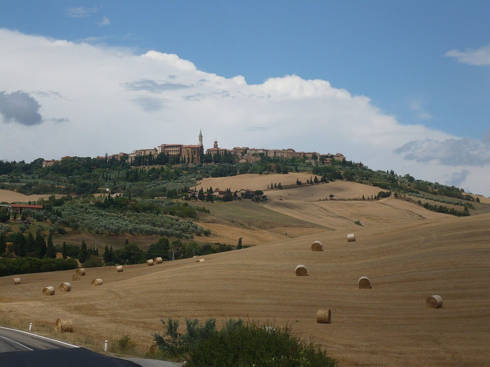 A hilltop town with a prominent tower, surrounded by golden fields with hay bales and green trees under a cloudy sky. A road is in the foreground.