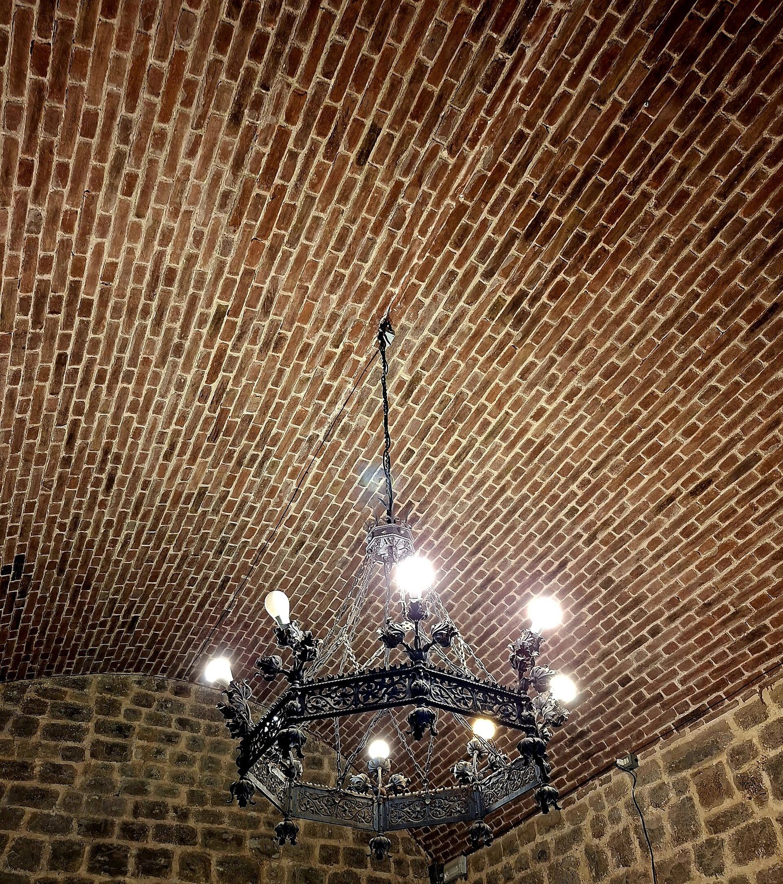 A vaulted ceiling constructed from reddish-brown bricks, with an ornate, dark metal chandelier featuring multiple illuminated bulbs hanging from its centre.