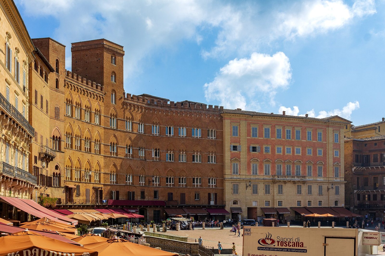 A wide view of Piazza del Campo in Siena, showing historic buildings, many outdoor cafe awnings, and people walking under a blue sky.