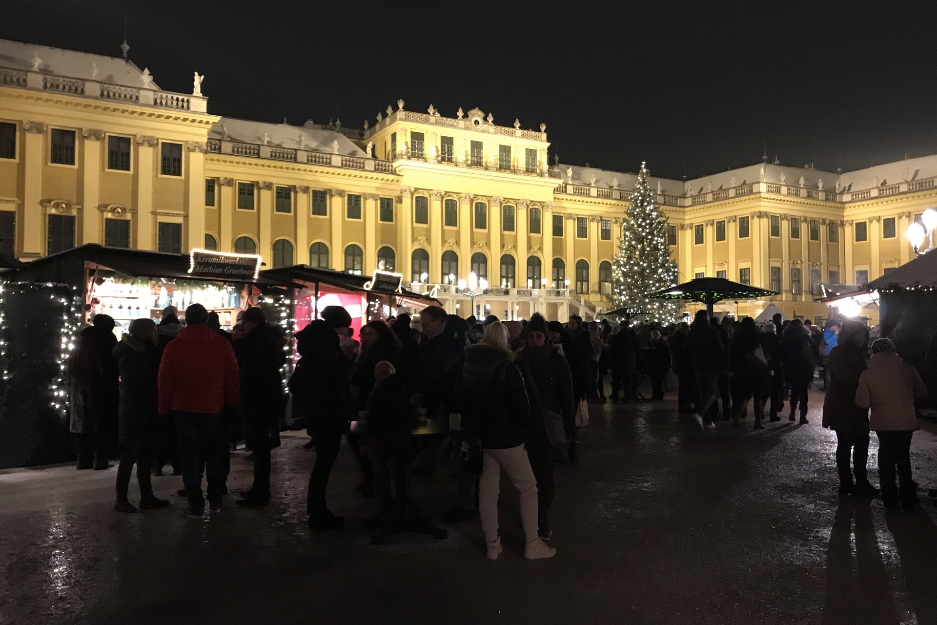 Crowds at the lit stalls of a Christmas market in front of the yellow facade of Schönbrunn Palace at night, with a large Christmas tree