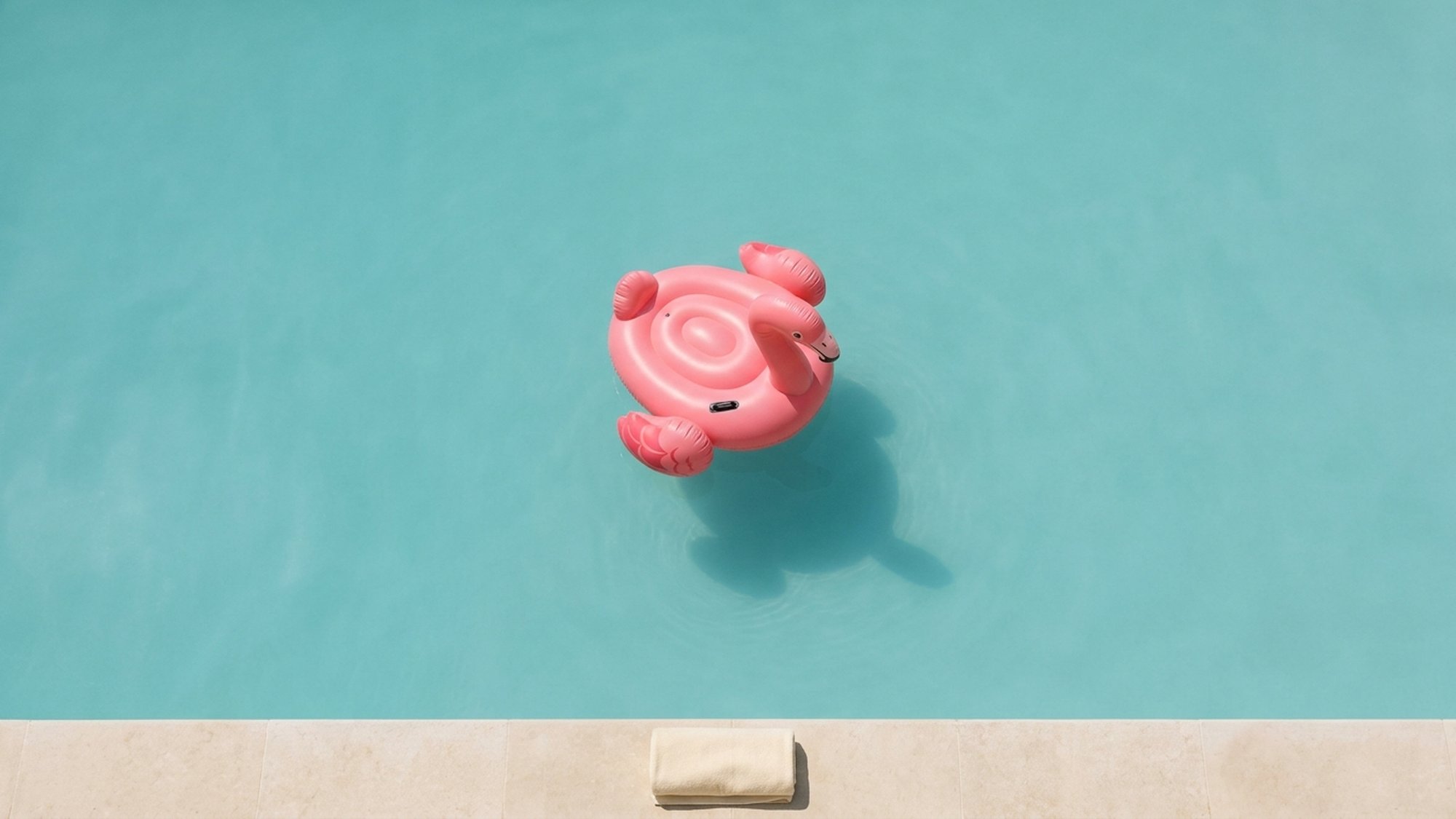 An overhead view of a single pink inflatable flamingo float alone in a turquoise swimming pool.