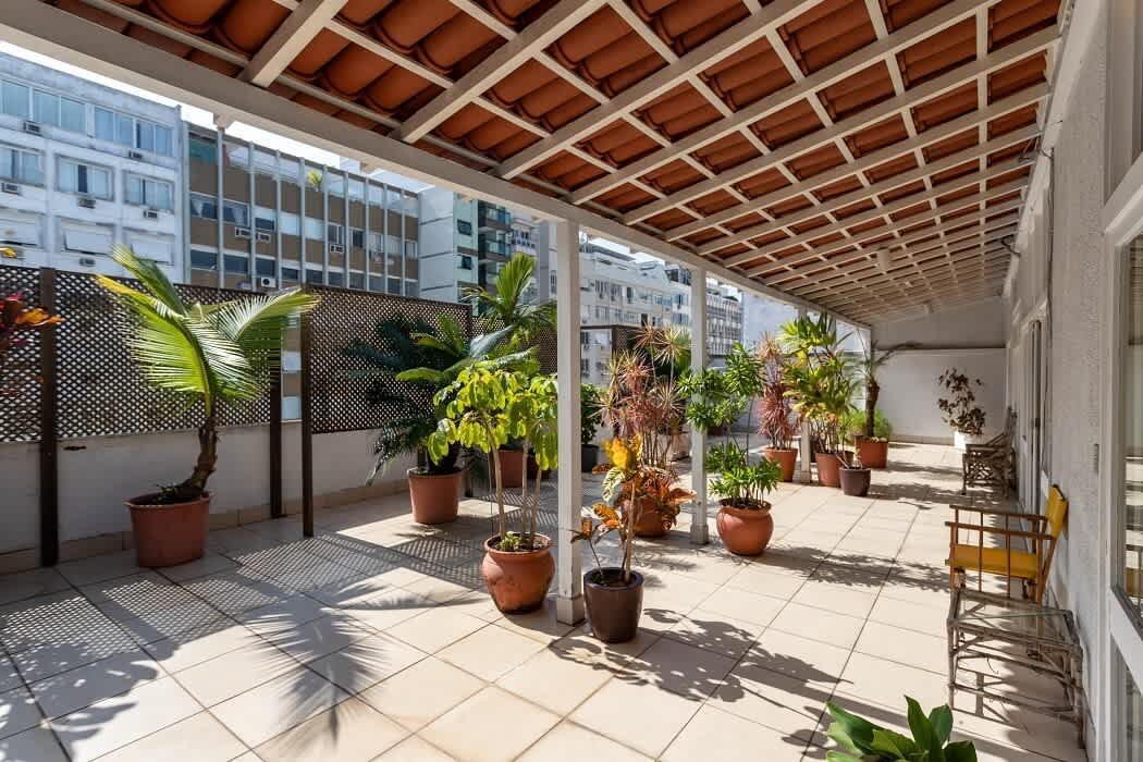 The covered wraparound terrace of the Ipanema Beach Linear Penthouse in Rio, with a terracotta-tiled pergola roof on white posts, lattice screens along the parapet, potted palms and plants in terracotta pots and a pale tiled floor