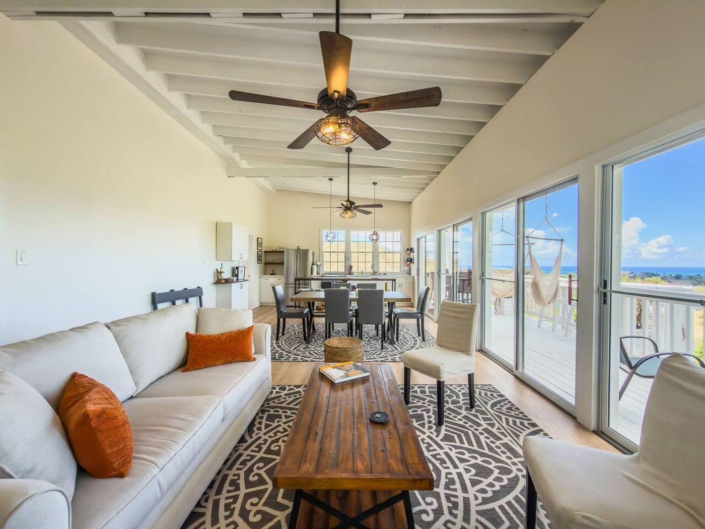 Living room with a white sofa, orange cushions and sliding glass doors along a long wall, a dining table beyond