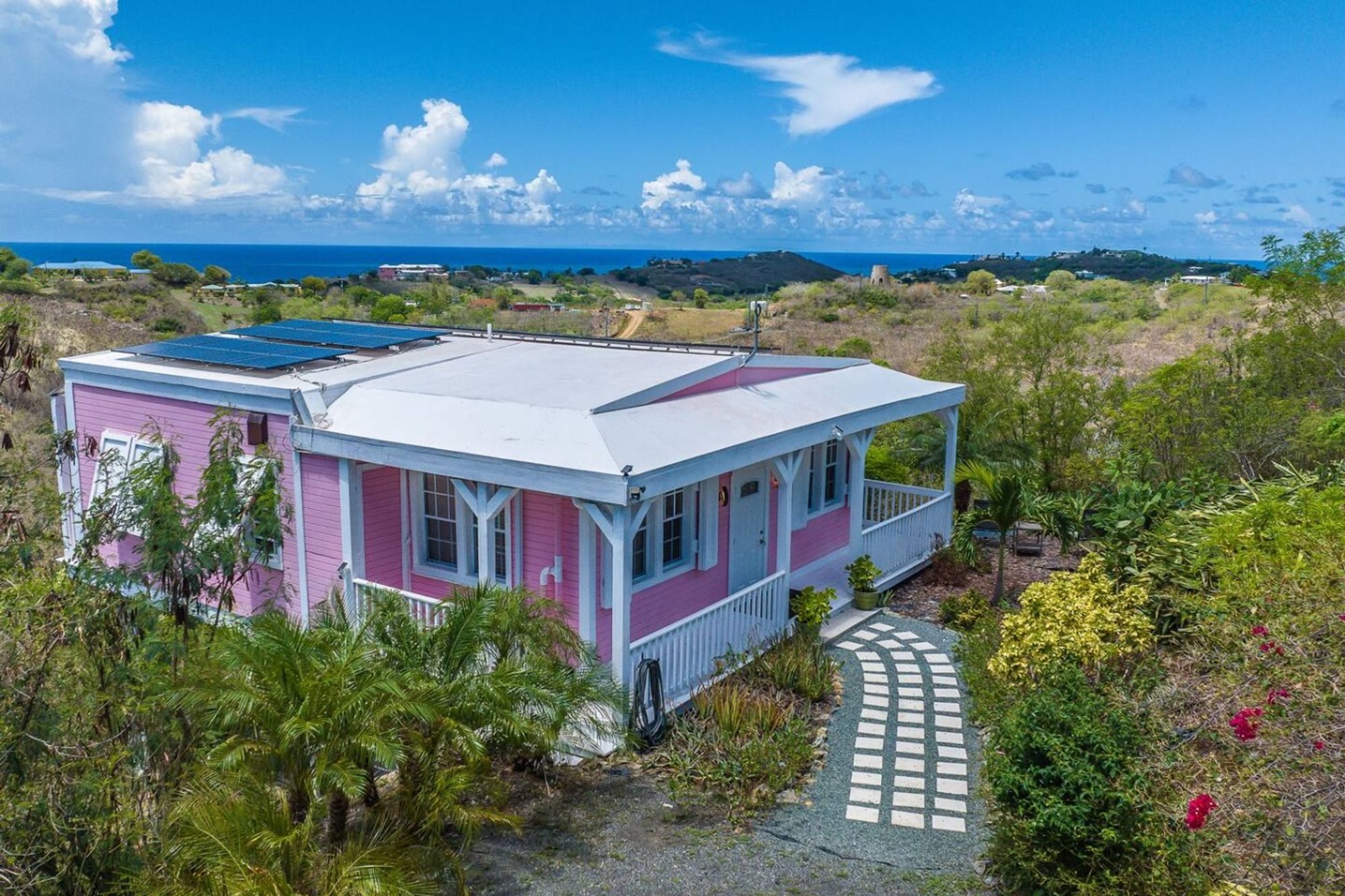 A pink cottage with a white roof and porch among tropical planting on a hillside, the sea on the horizon