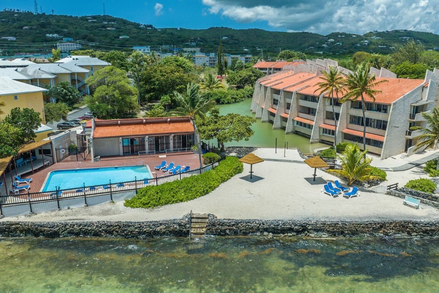 Aerial view of the complex's pool, loungers and palm trees beside the beach and shallow turquoise water