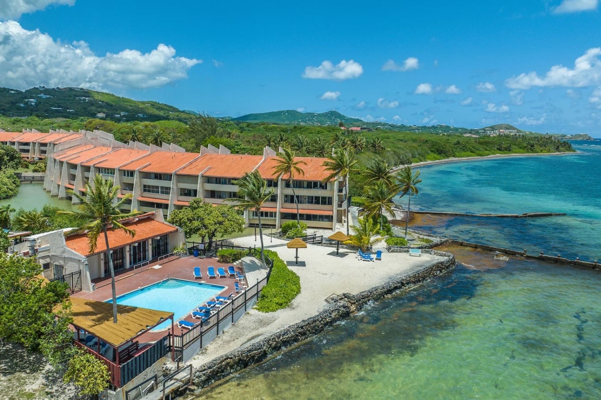 Aerial view of a low terracotta-roofed condominium complex on the water, a pool and loungers beside a small beach and a reef-lined bay