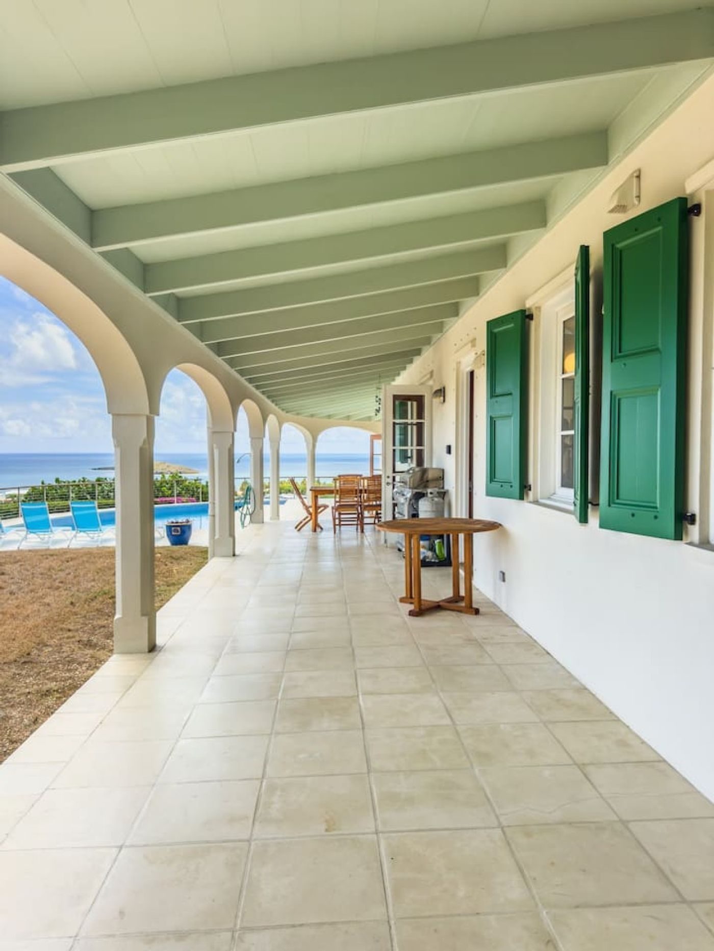 Long covered veranda with white arches, a tiled floor and green shutters, open to the sea at the far end