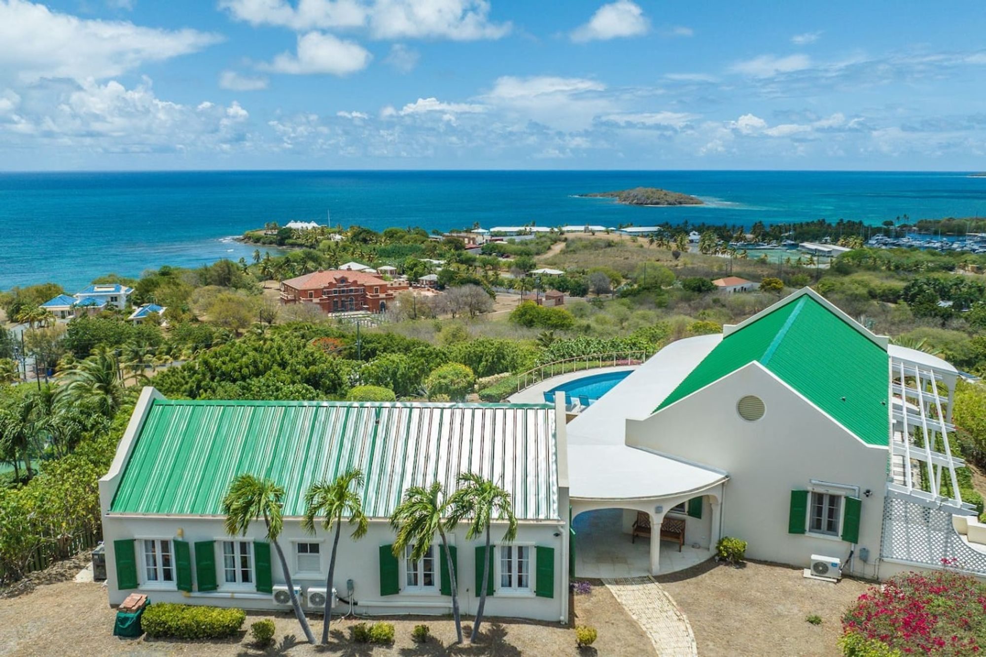A white villa with green metal roofs and green shutters on a hillside, with the sea and small offshore islands behind