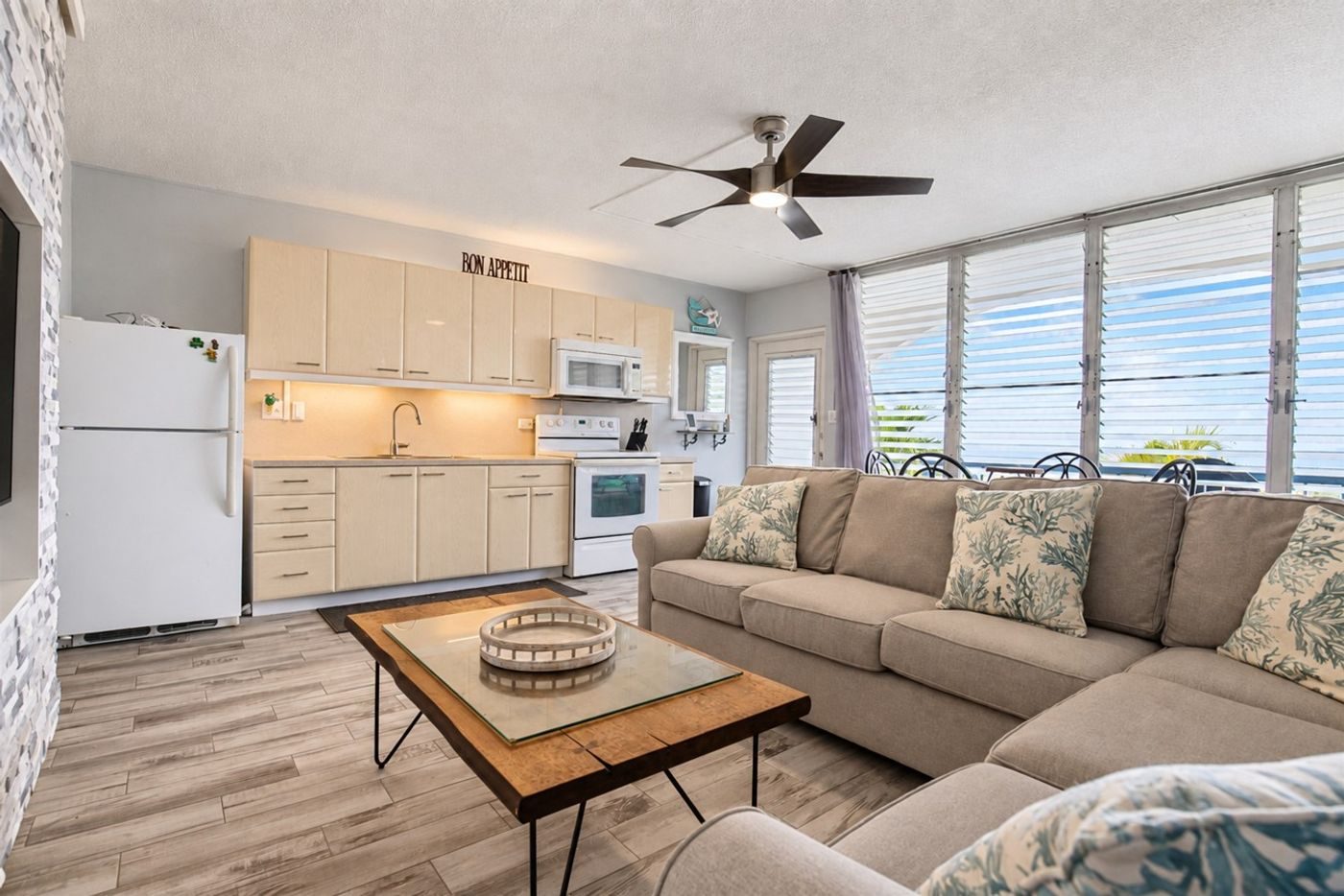 Living room and kitchen in one bright room with louvred windows along one wall and a ceiling fan